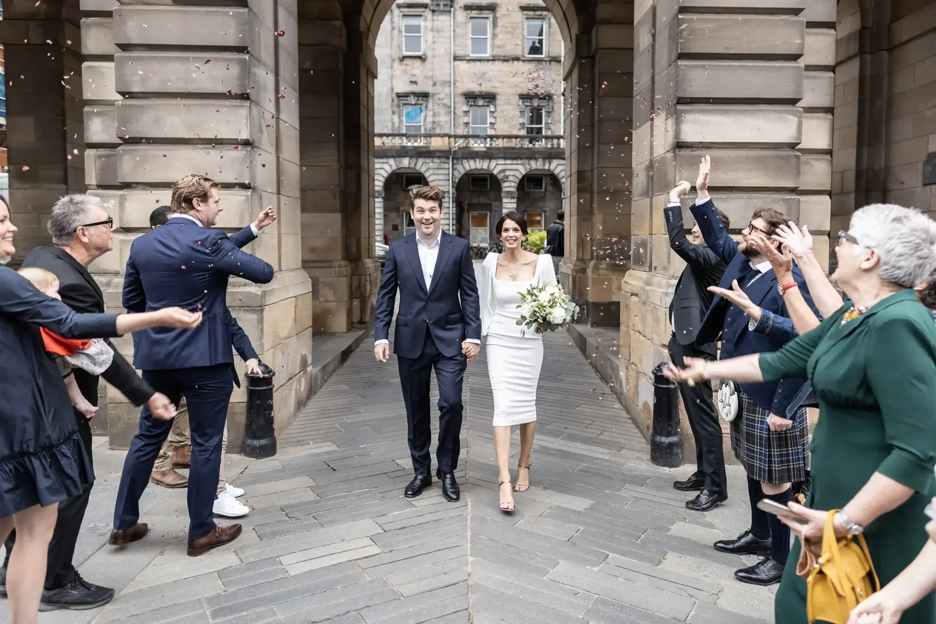 A newlywed couple walks through a joyous crowd throwing confetti, with a historic building in the background.