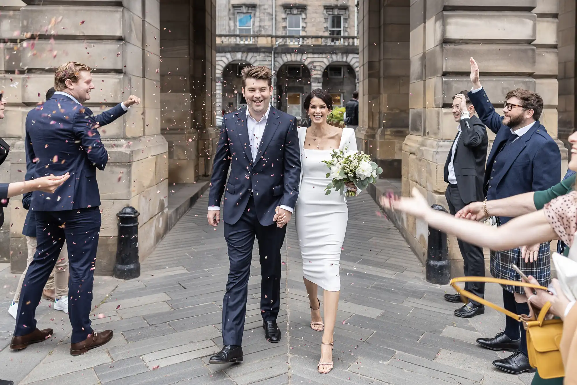 A newlywed couple walking joyfully through a crowd as guests throw confetti, celebrating outside a stone building.