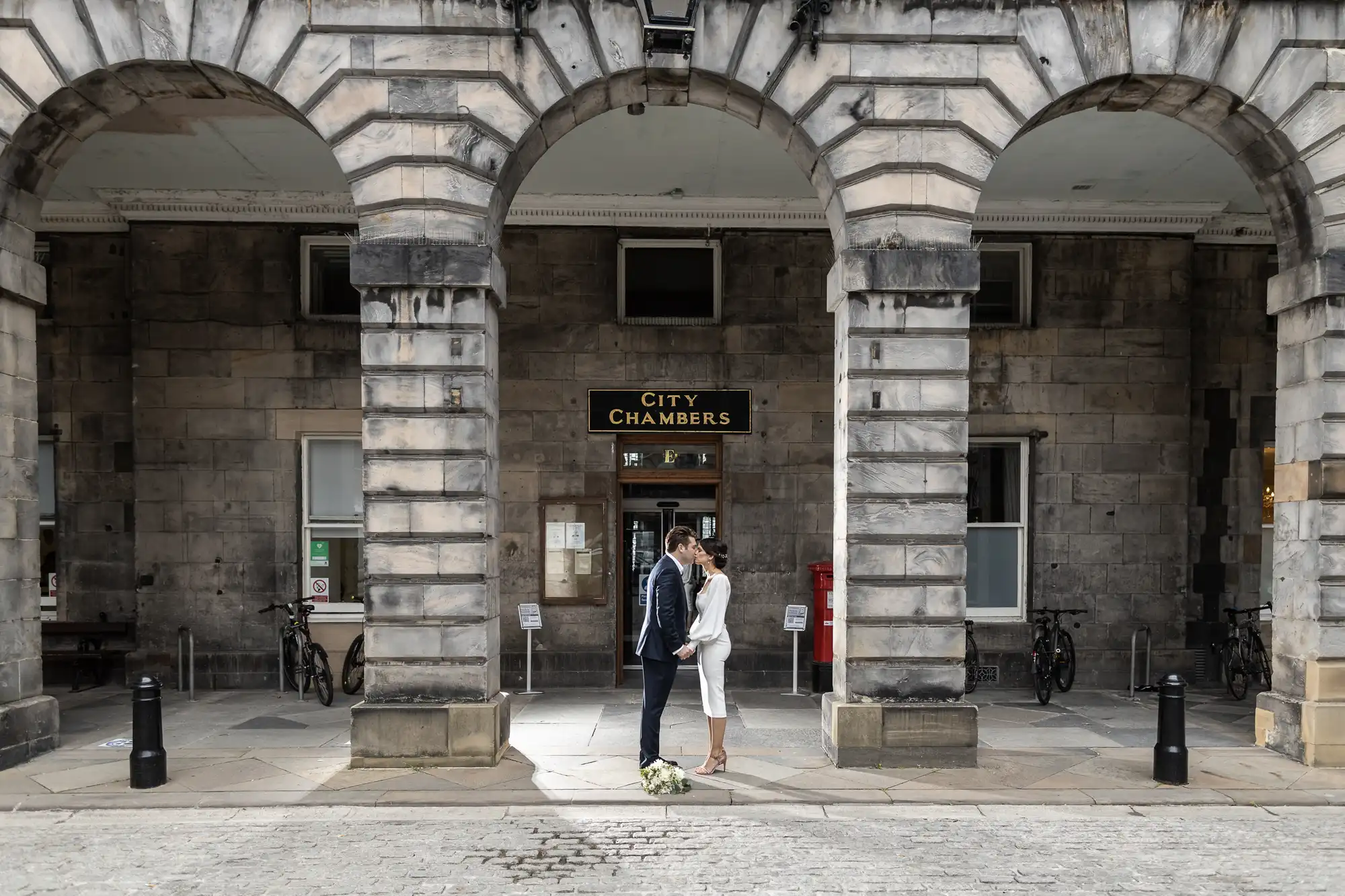 A couple kisses under the archway of the city chambers building, with bicycles parked nearby and the structure exhibiting grand stonework.