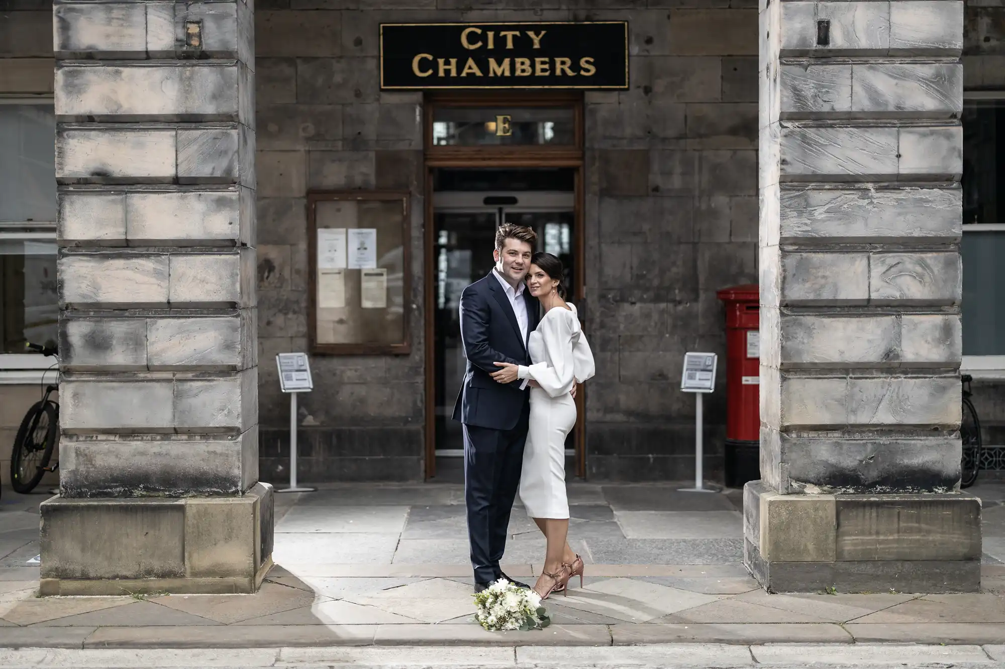 A couple embracing and smiling outside the city chambers, with the bride in a white suit and the groom in a dark suit, standing next to a bouquet on the ground.