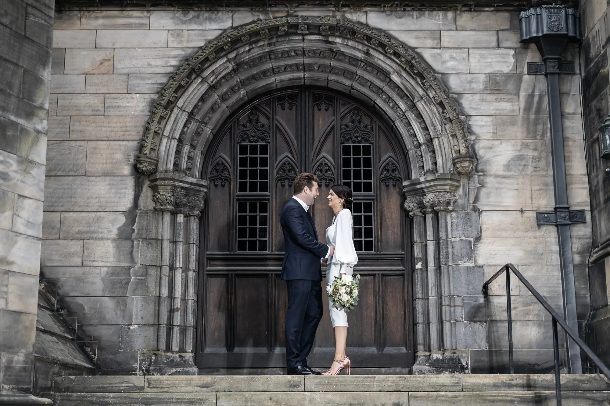 A couple kissing on steps in front of a church's ornate gothic doorway, the bride holding a bouquet and the groom in a suit.