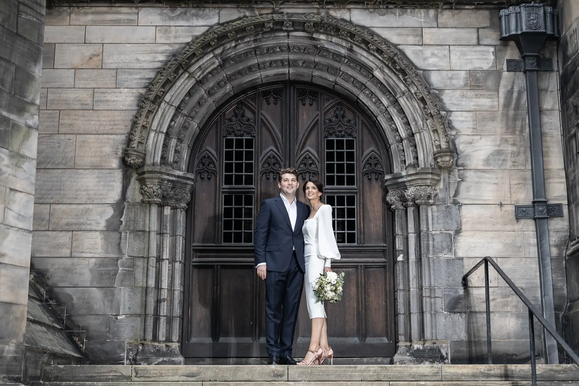 A newlywed couple stands smiling in front of an ornate church doorway, the bride holding a bouquet and both dressed in elegant wedding attire.
