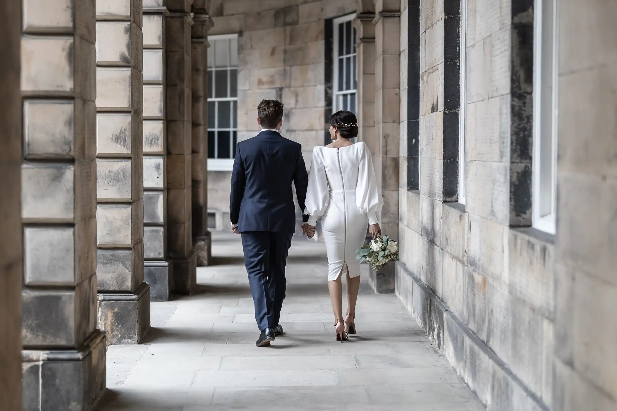 A newlywed couple walks hand in hand along a stone-columned corridor, the woman in a white dress carrying a bouquet and the man in a navy suit.