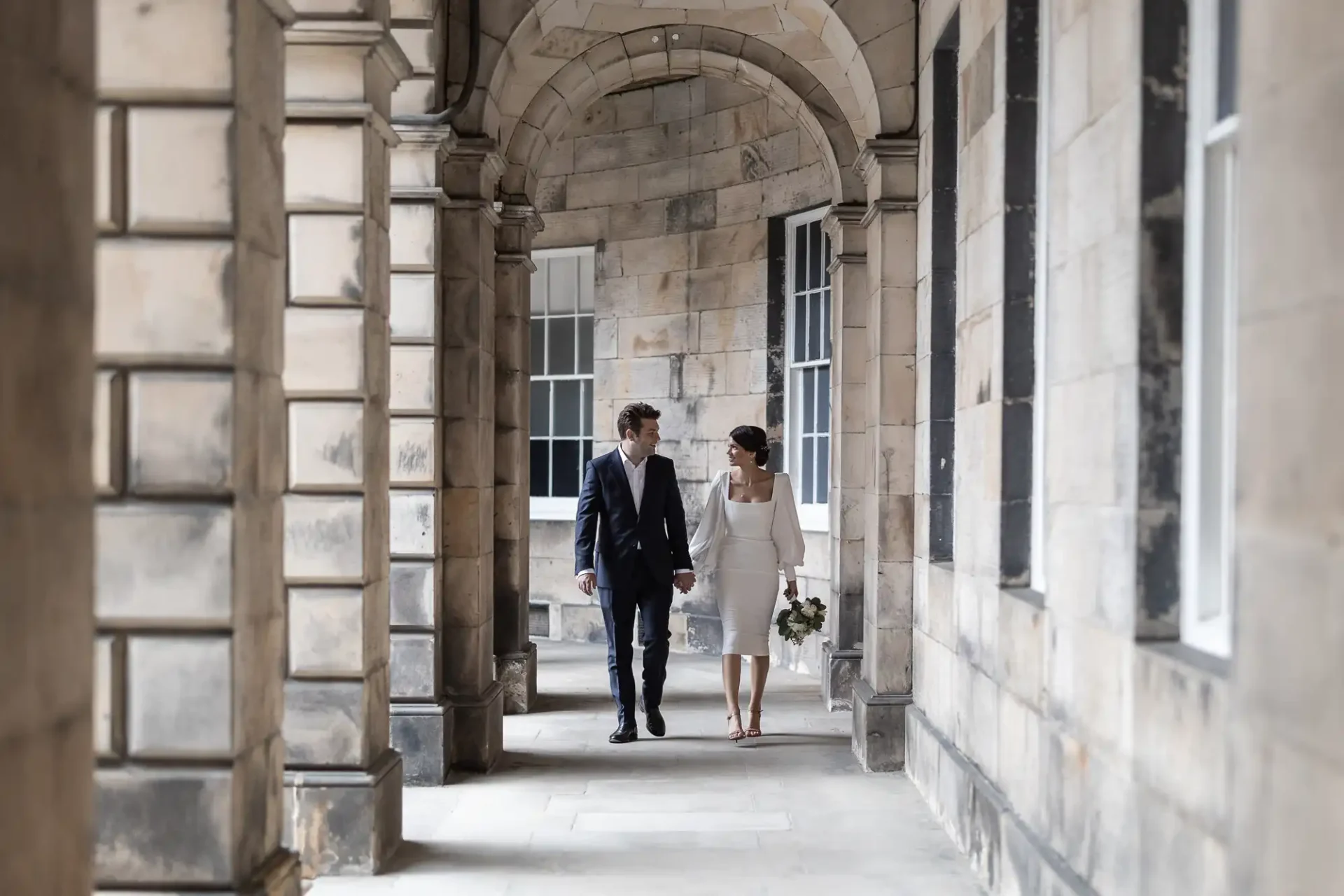 A bride and groom holding hands and walking through a stone arcade with archways.