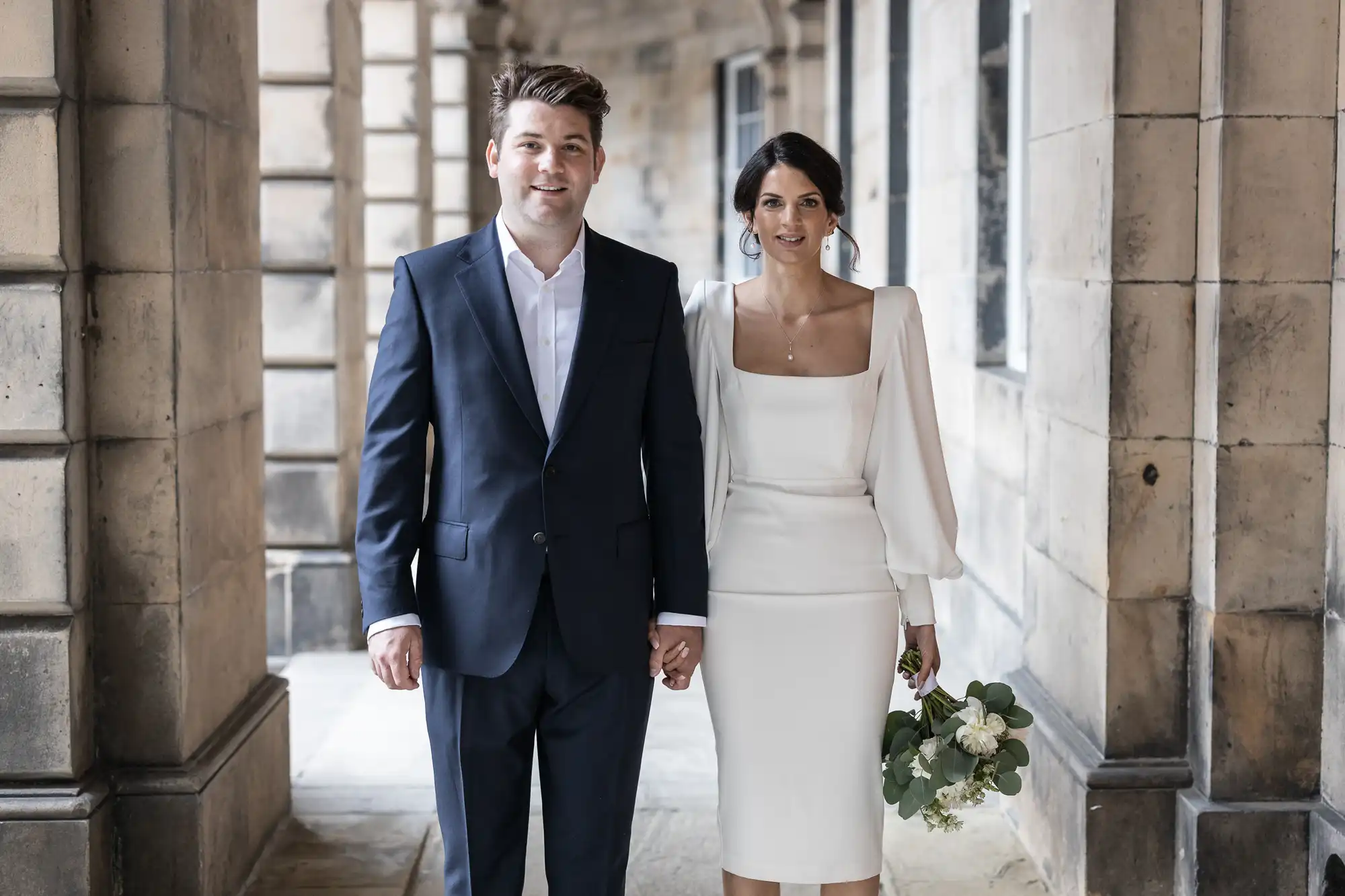 A newlywed couple walking hand in hand through a stone-pillared corridor, the bride in a white dress and carrying a bouquet, and the groom in a navy suit.