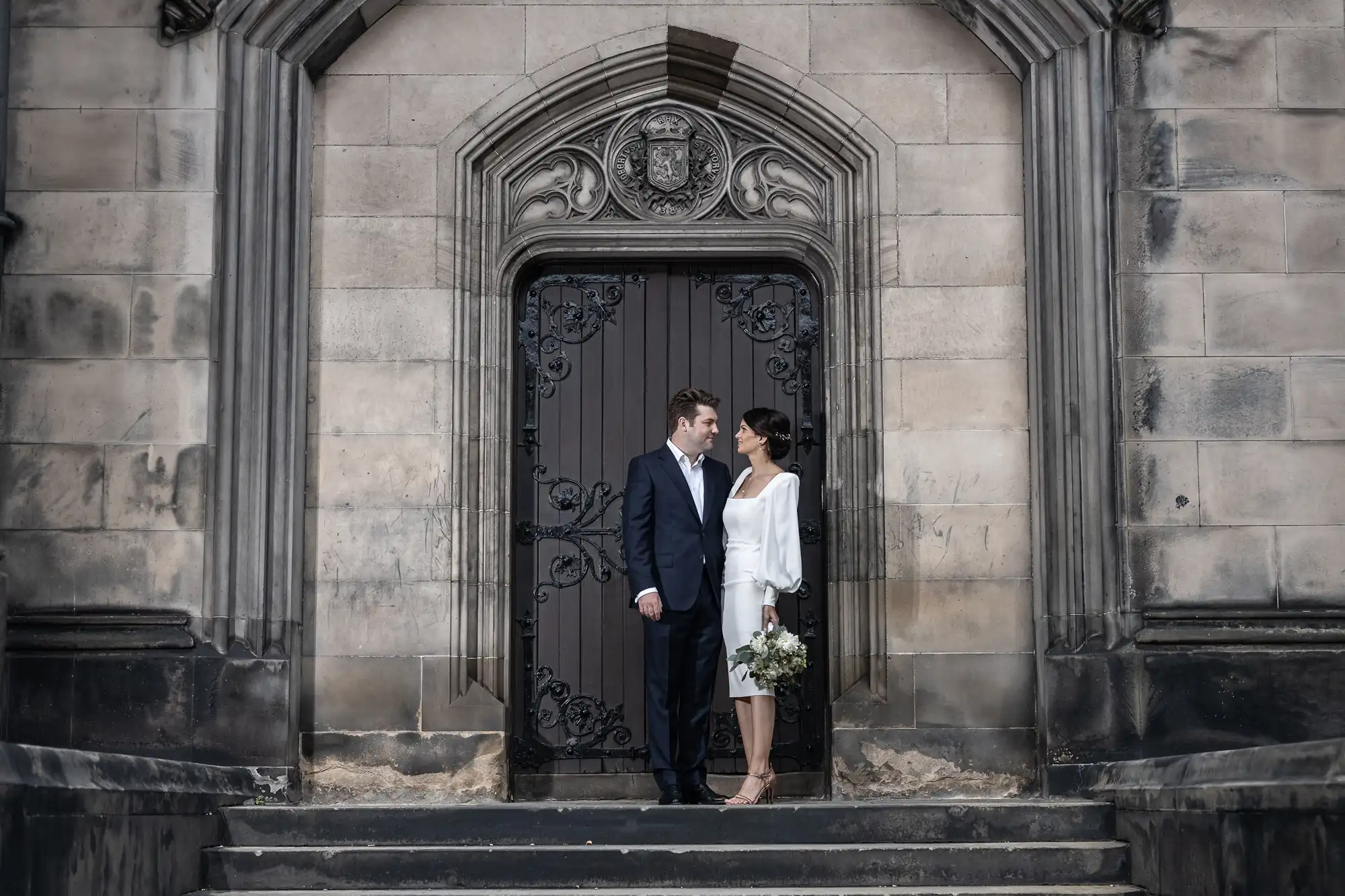 A couple stands closely on the steps of an ornate, historic doorway, exchanging smiles, with the woman holding a bouquet.