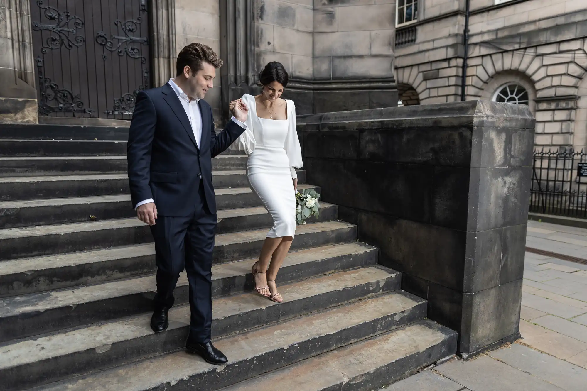A man in a suit and a woman in a white dress walk down steps outside a building, the woman holding her shoes.