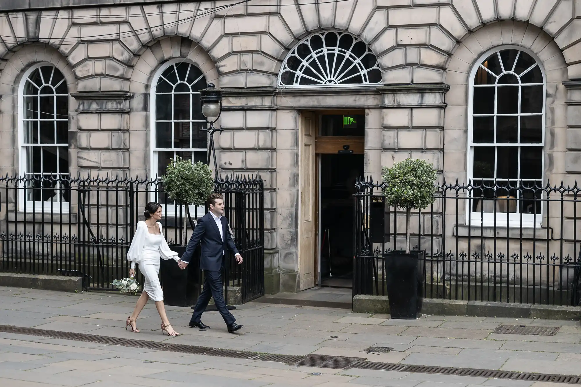A couple dressed smartly, the woman in a white dress and the man in a suit, walking past an elegant building with arched doorways.