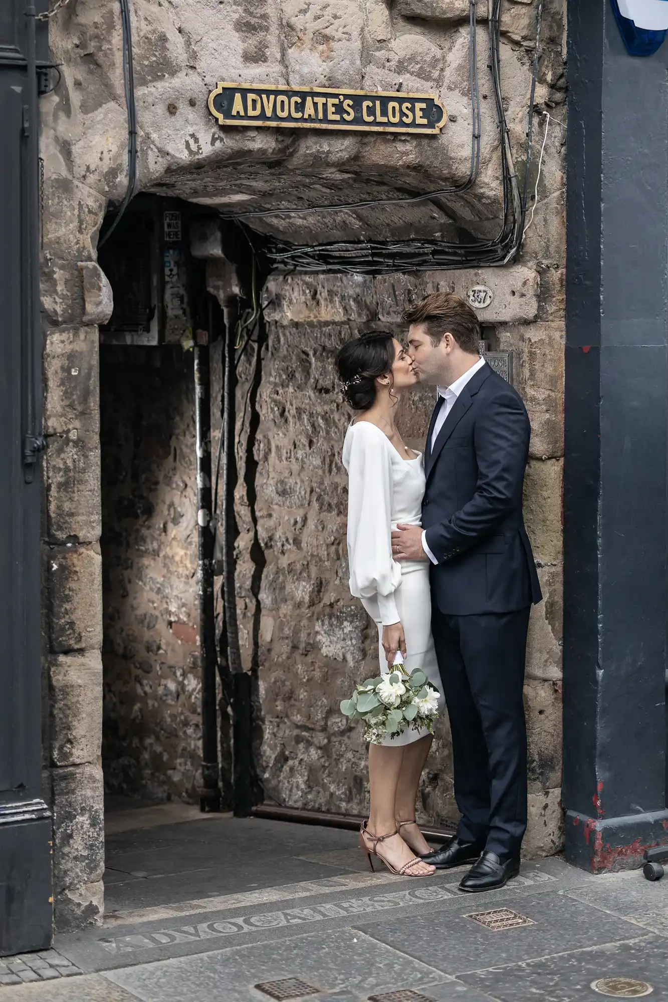 A couple kissing at the entrance of advocate's close, edinburgh, with the woman holding a bouquet and the man in a suit.