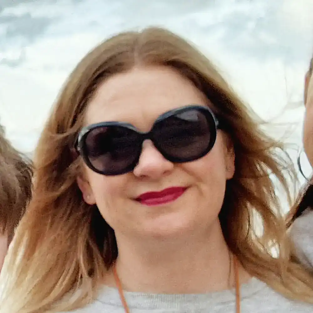 Person wearing black sunglasses and smiling, with long hair and a neutral background.