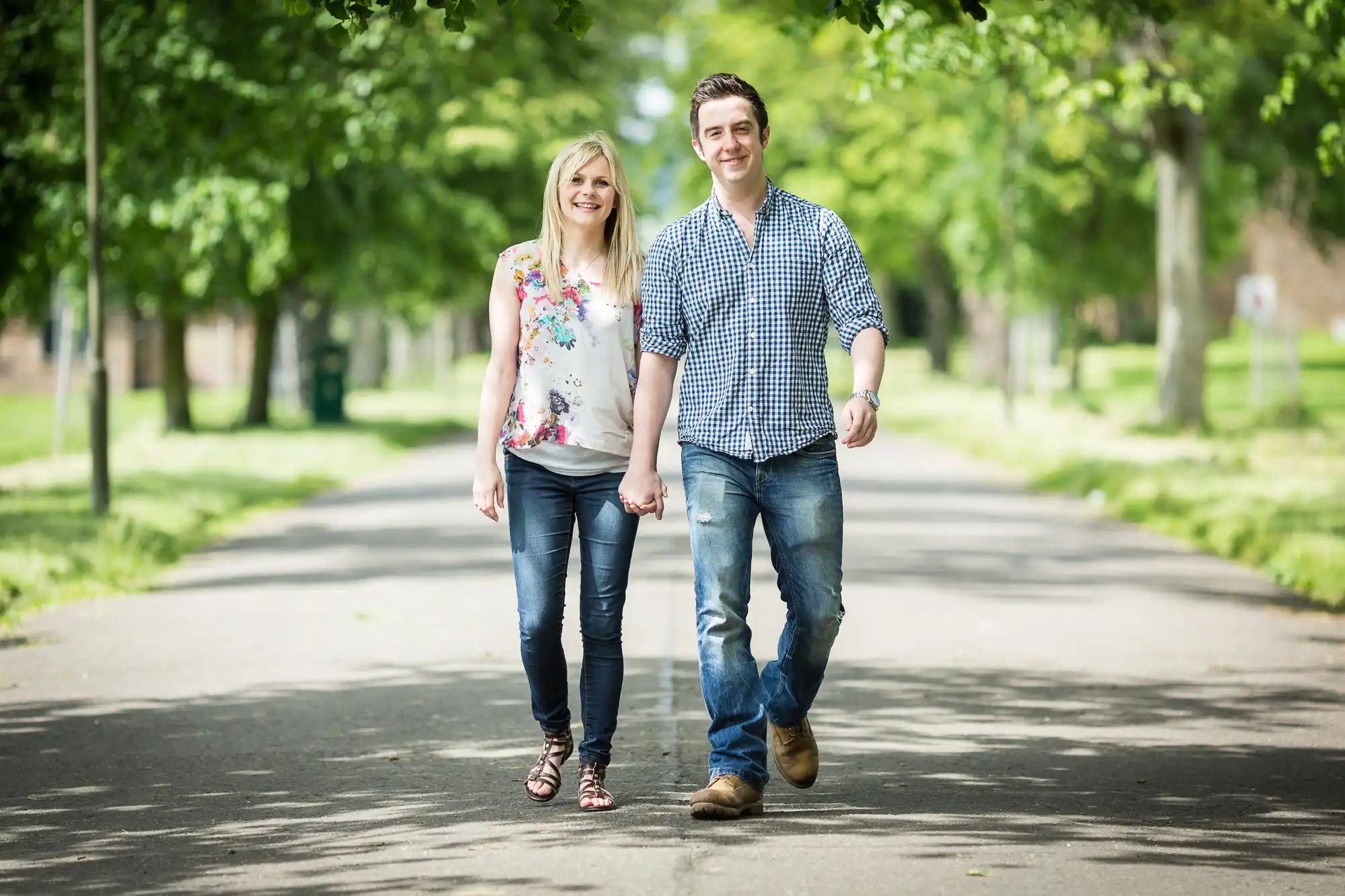 A young couple holding hands while walking down a tree-lined path on a sunny day.