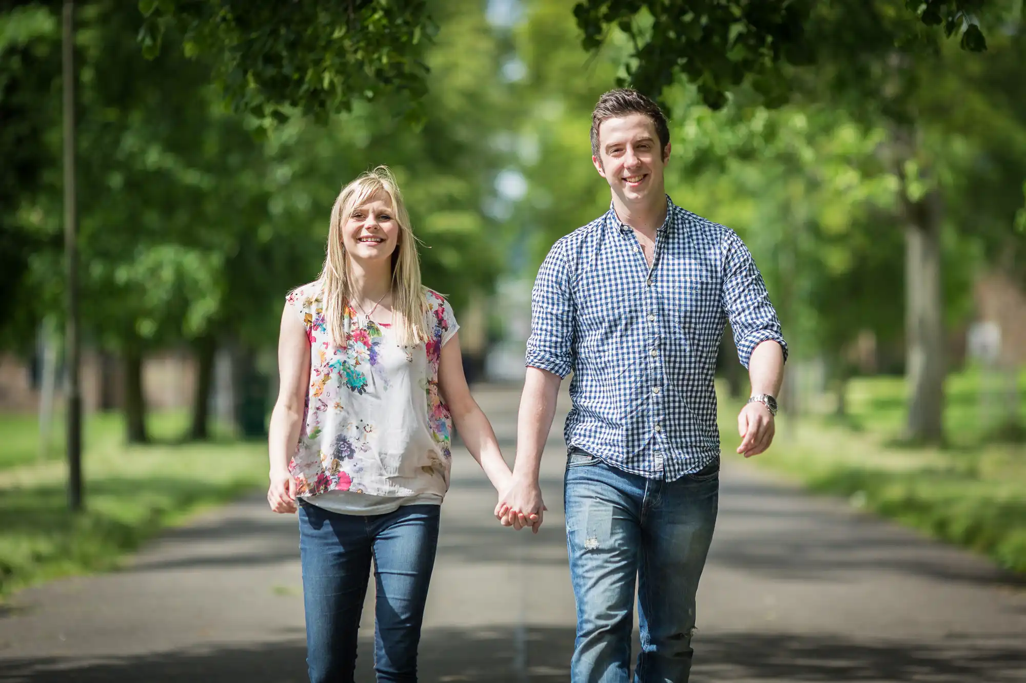 A young man and woman holding hands and smiling while walking down a tree-lined path in a park.