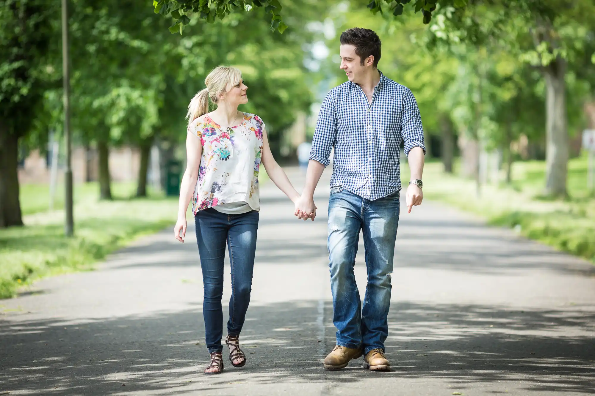 A young couple holding hands while walking down a tree-lined path in a park.