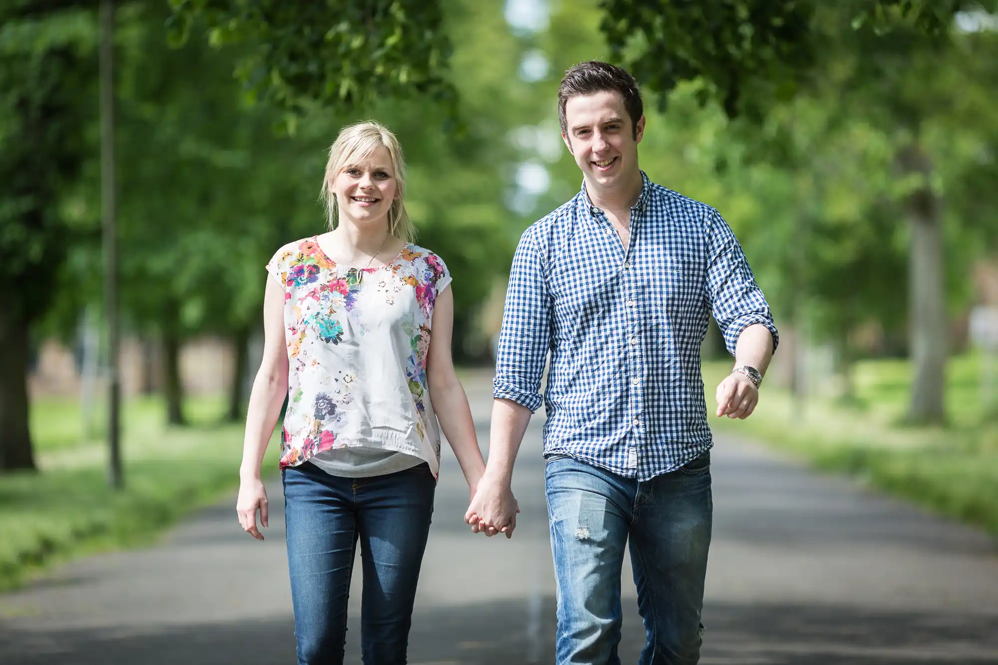 A man and a woman holding hands while walking on a tree-lined path, both smiling and looking towards the camera.