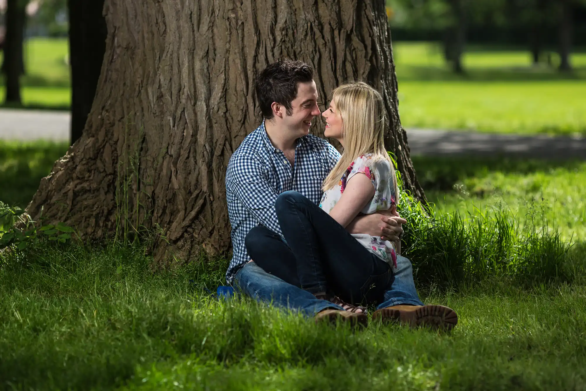 A couple sitting and smiling at each other by a large tree in a lush green park.