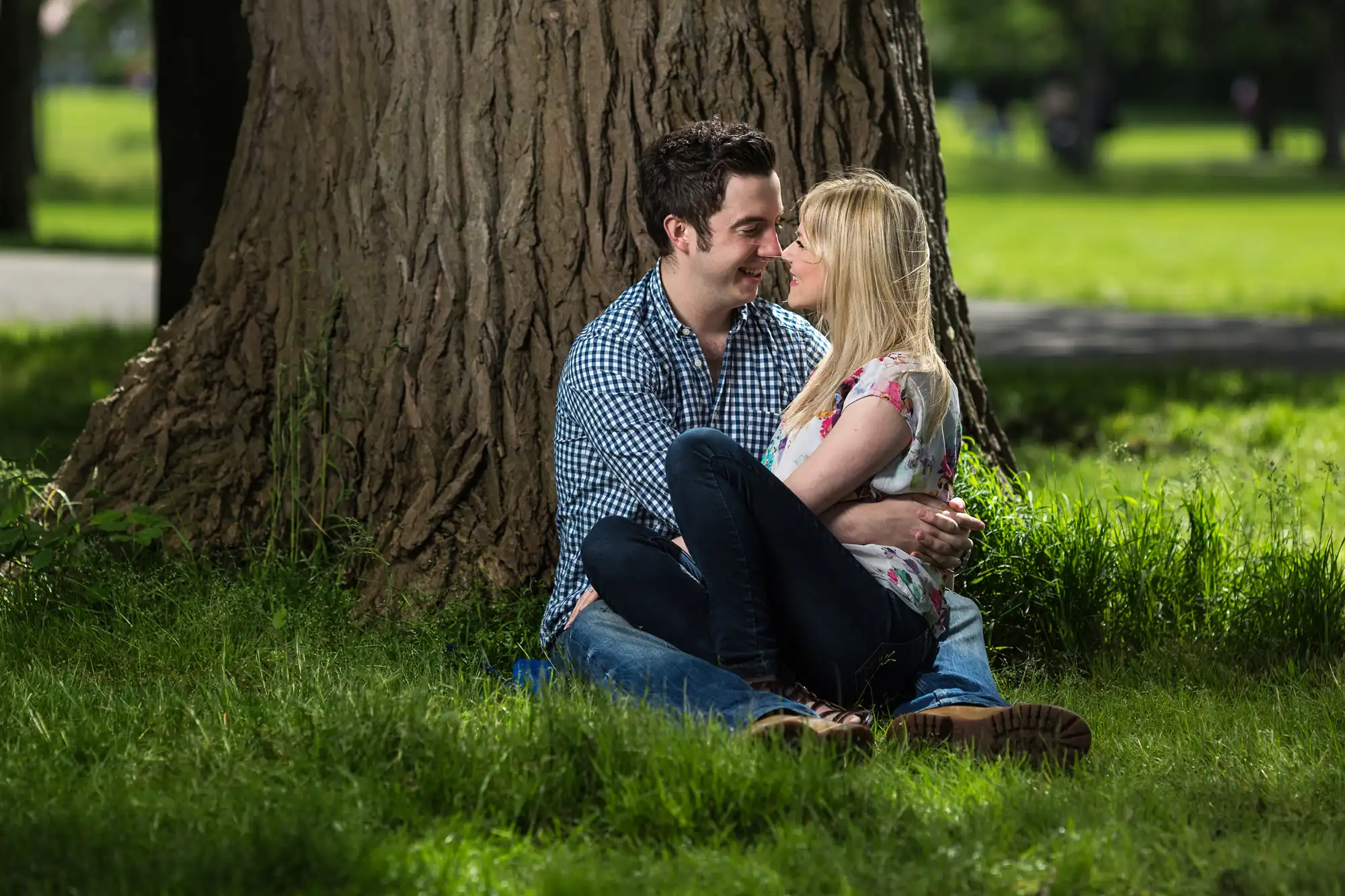 A couple sitting closely under a tree in a park, smiling at each other, surrounded by lush green grass.