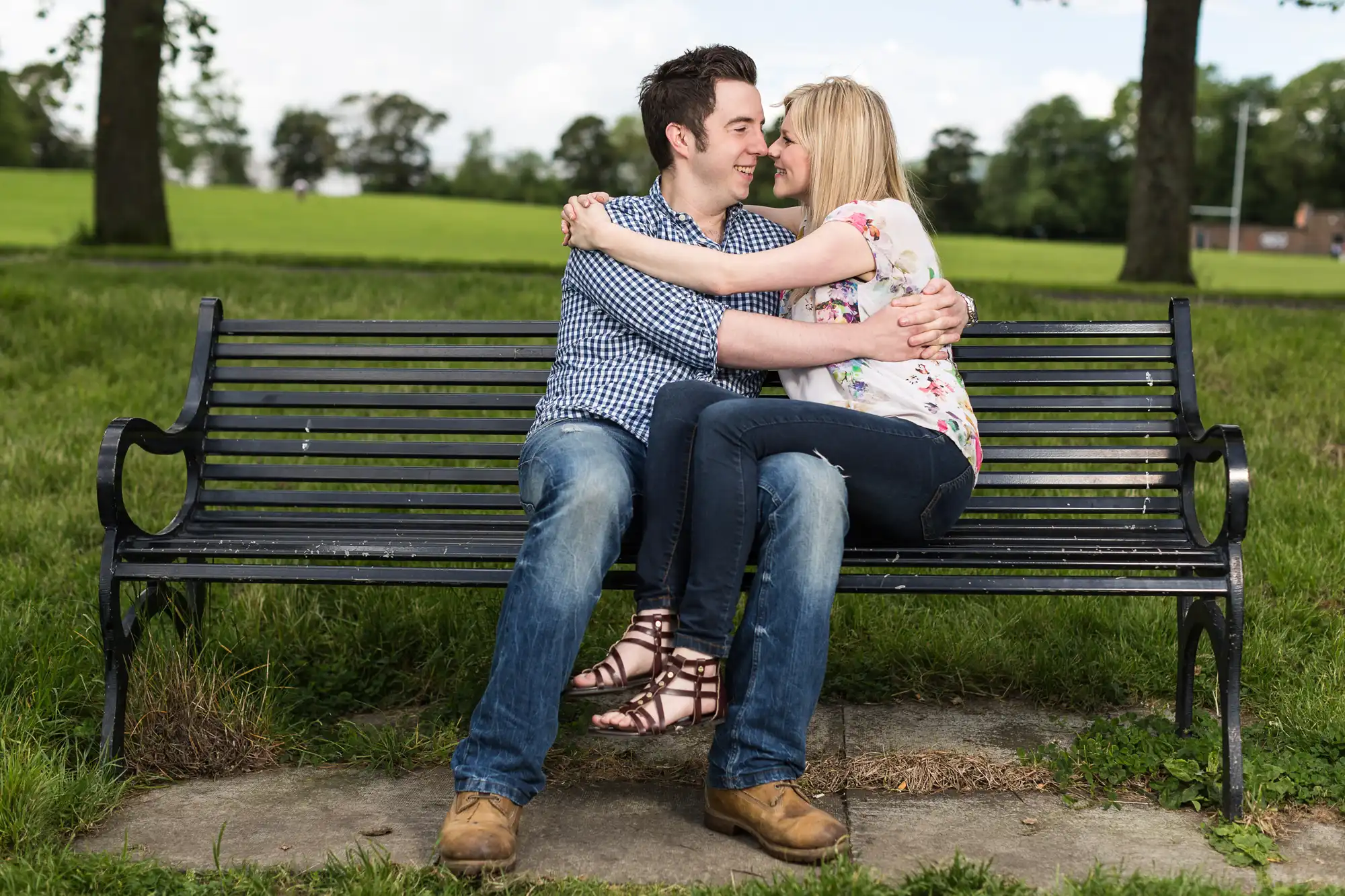A young couple sitting closely on a park bench, embracing and smiling at each other, with trees and grass in the background.
