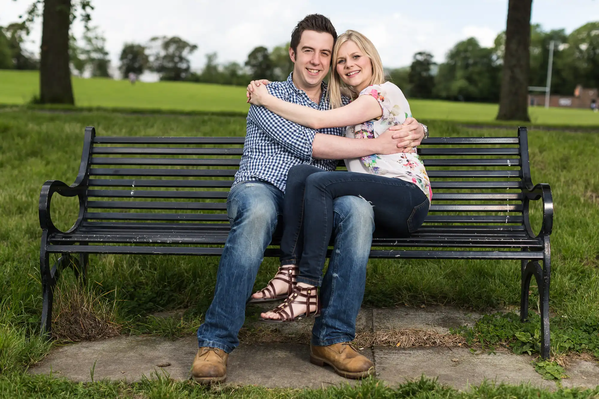 A young couple smiling and embracing on a park bench in a grassy area.