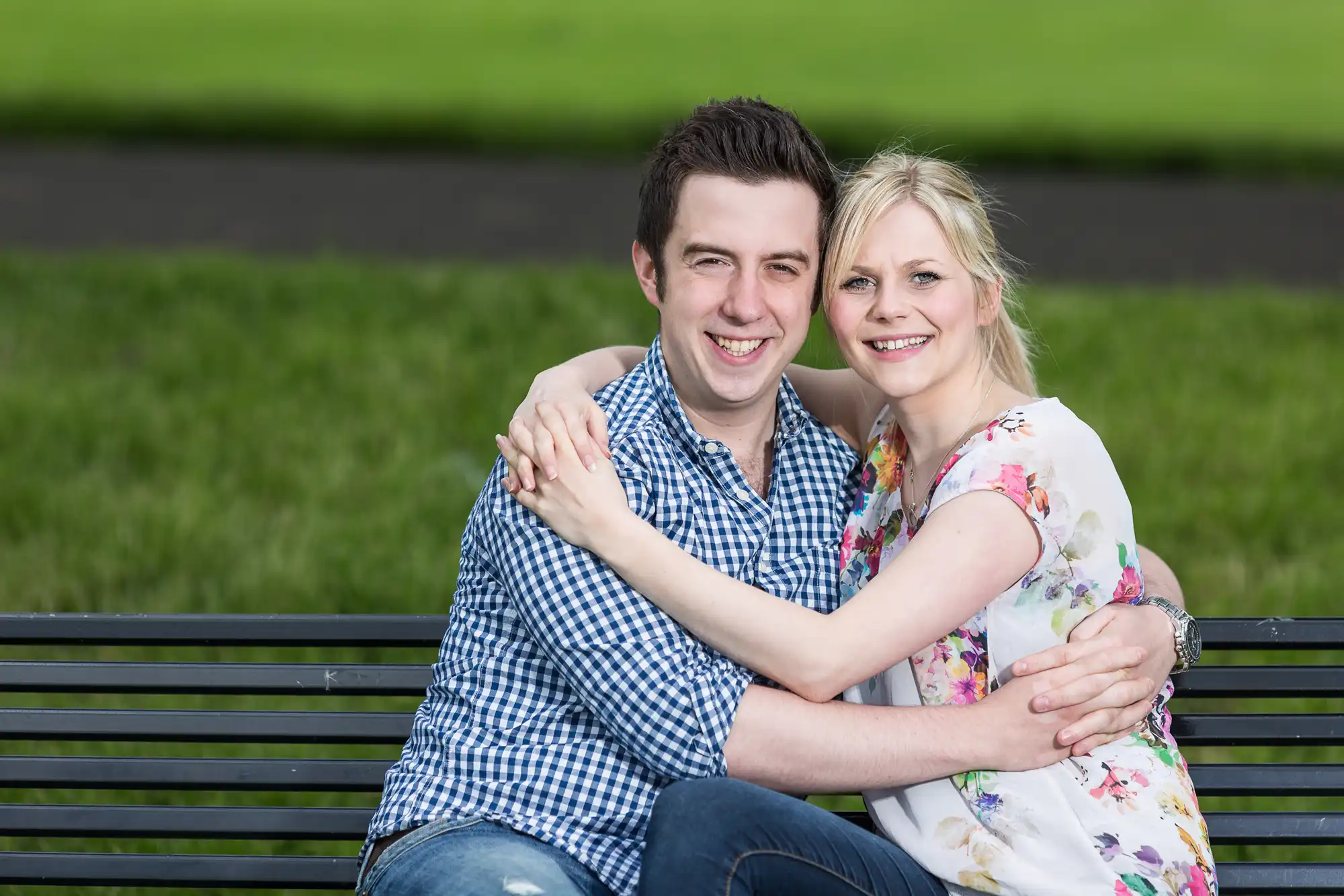 A smiling couple sitting closely together on a park bench, embracing each other against a blurred green grass background.