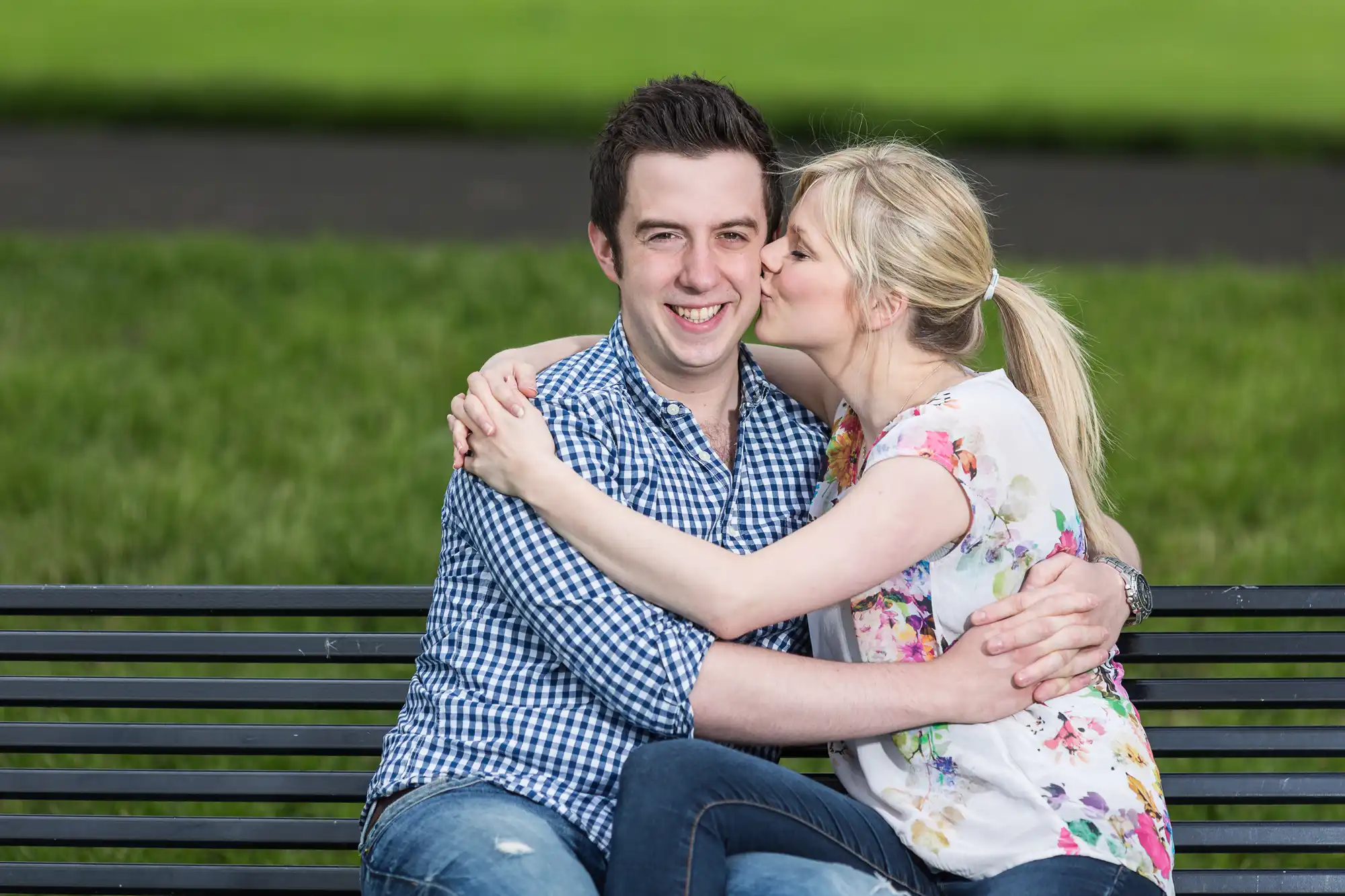 A woman kissing a smiling man on the cheek as they sit together on a park bench, with a grassy background.