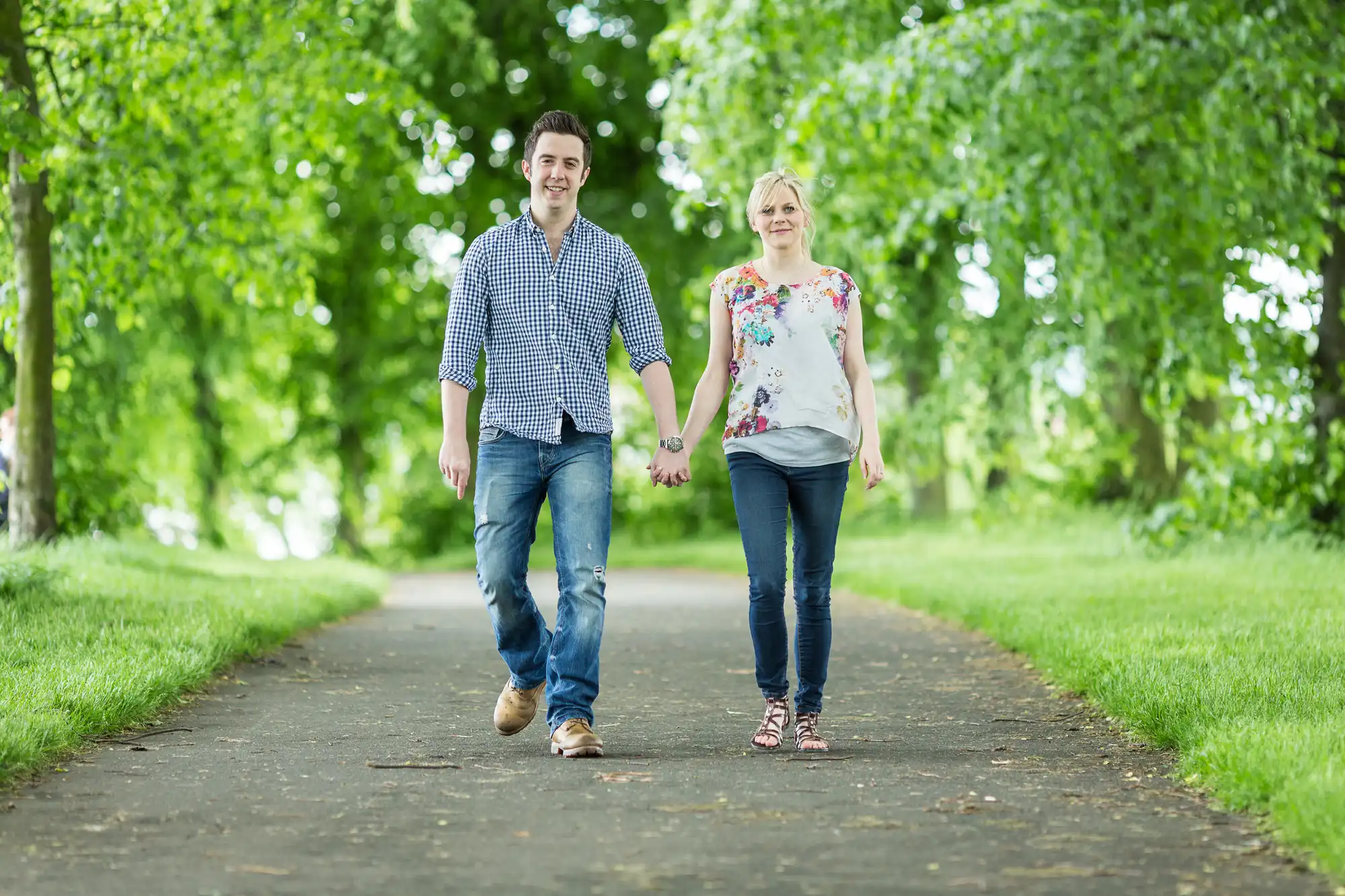 A young couple holding hands while walking down a tree-lined pathway in a park.