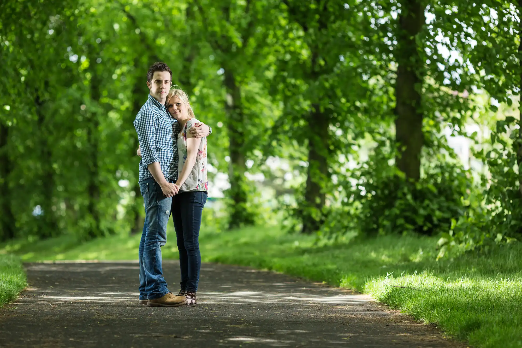 A couple embracing each other on a tree-lined path, with the man standing behind the woman, both looking gently towards the camera.