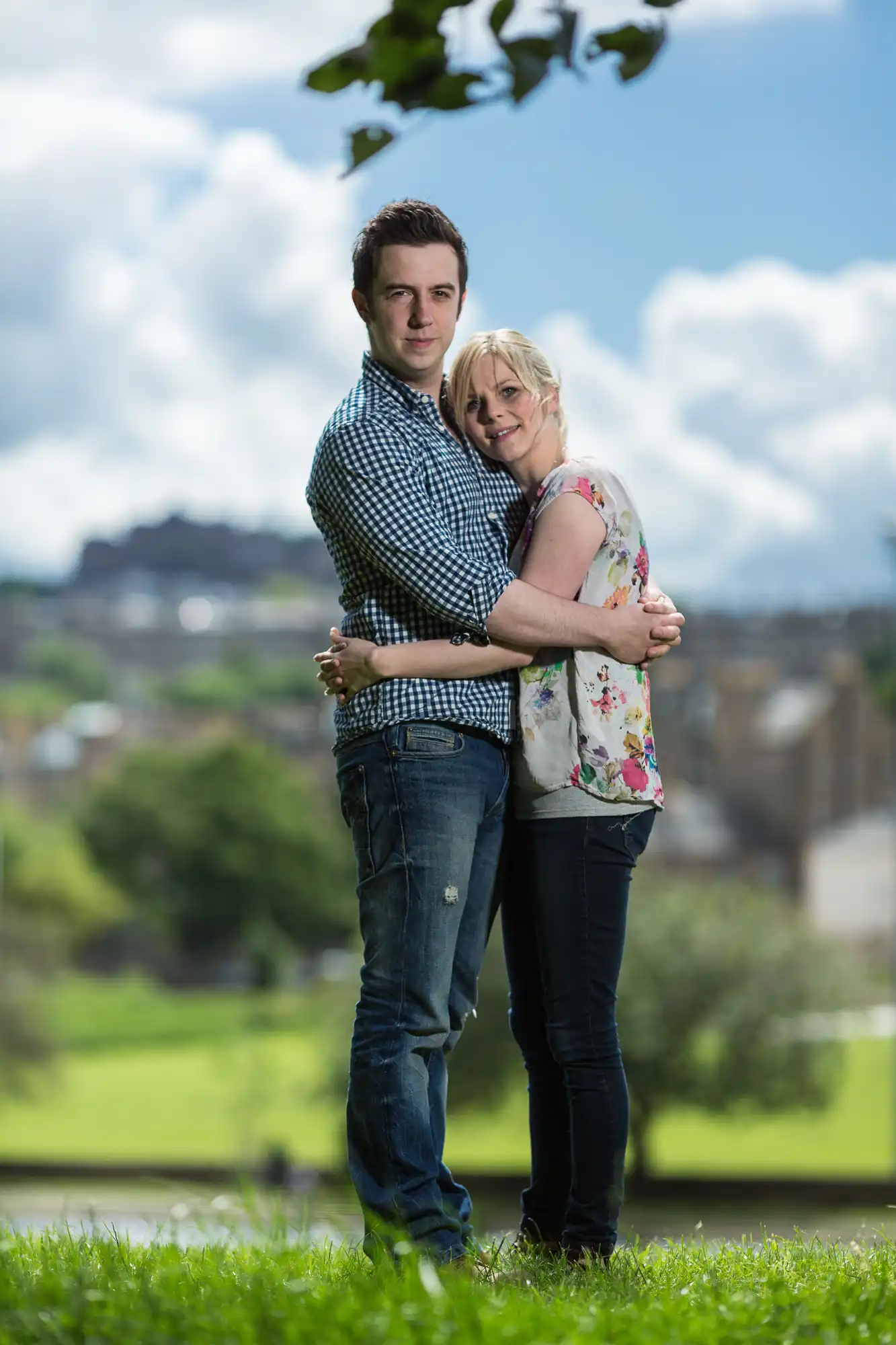 A young man and woman embracing in a sunny park, with a cityscape in the blurred background.