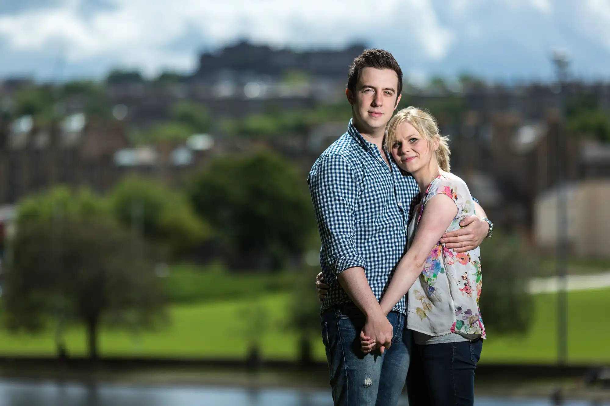 A young couple embracing outdoors with a blurry cityscape and lush greenery in the background.