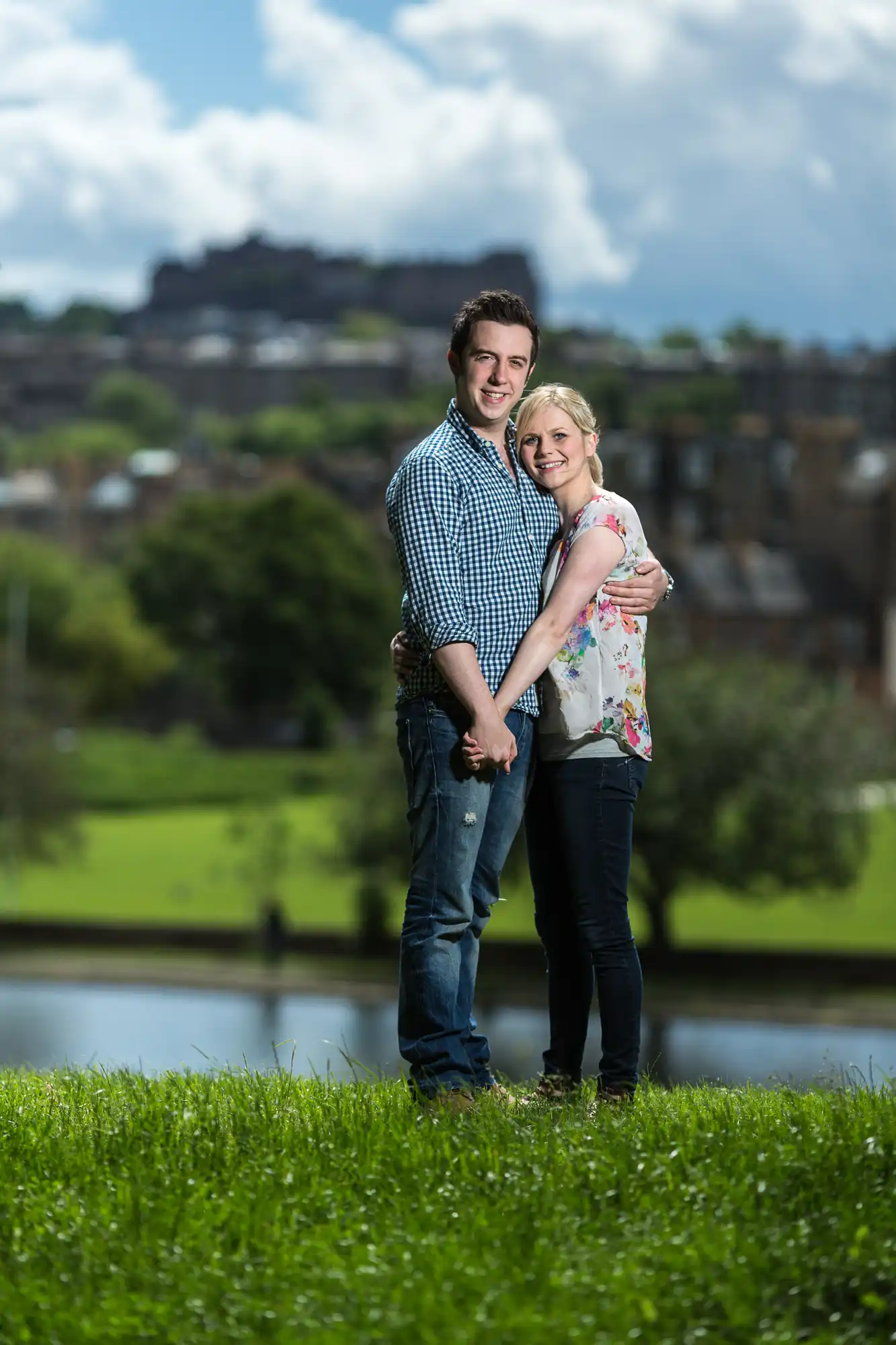 A couple embracing in a grassy field with a cityscape and castle in the background on a sunny day.