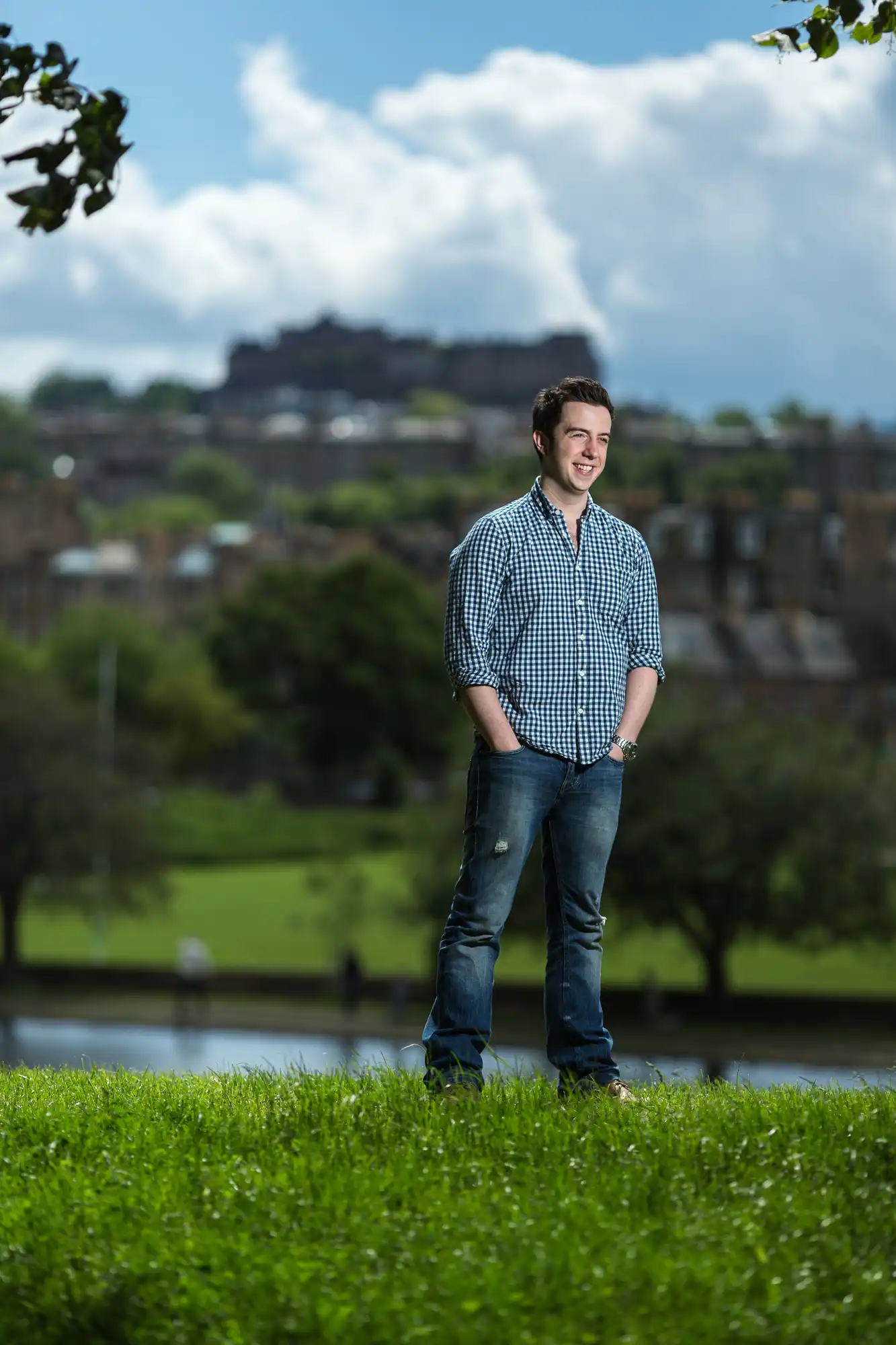 A young man in a checked shirt and jeans stands smiling in a park, with a castle on a hill visible in the background.