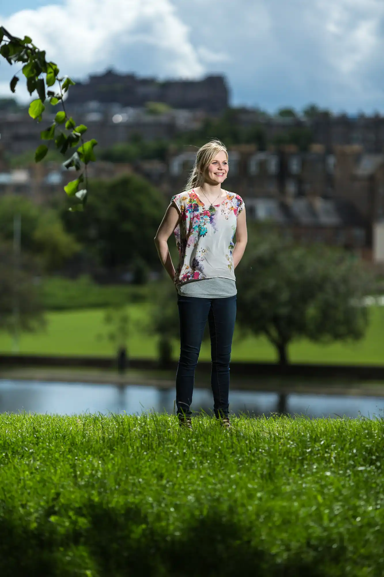 Woman standing on grass with a scenic cityscape in the background, wearing a floral top and jeans, smiling.