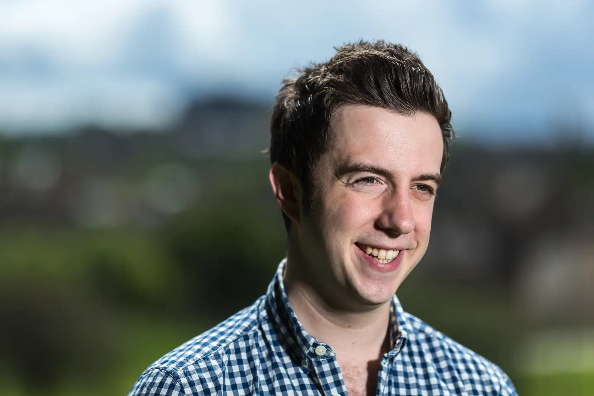 Smiling man in a blue checked shirt with a blurred natural background.