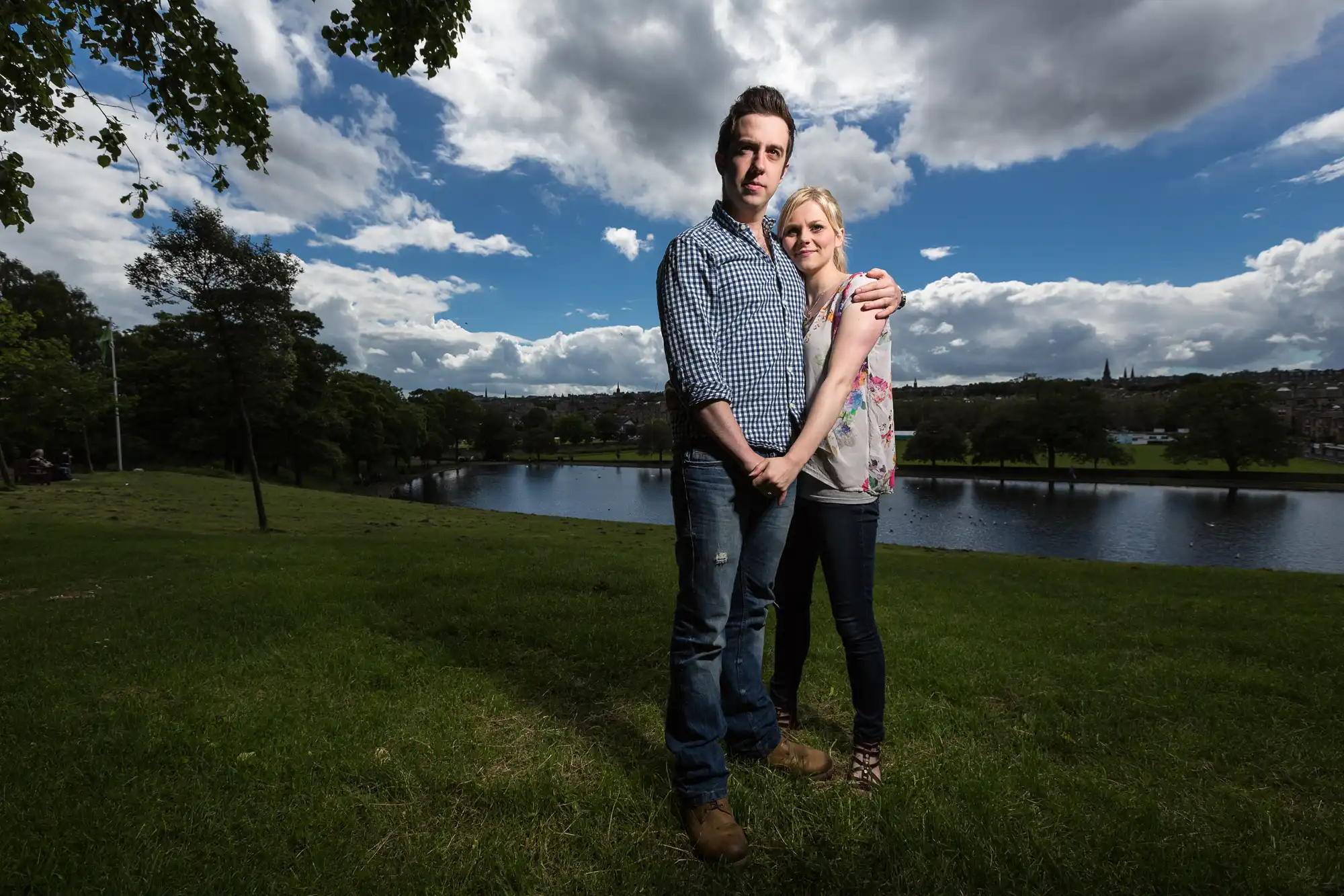 A couple embracing in a sunny park with a lake and distant cityscape in the background.