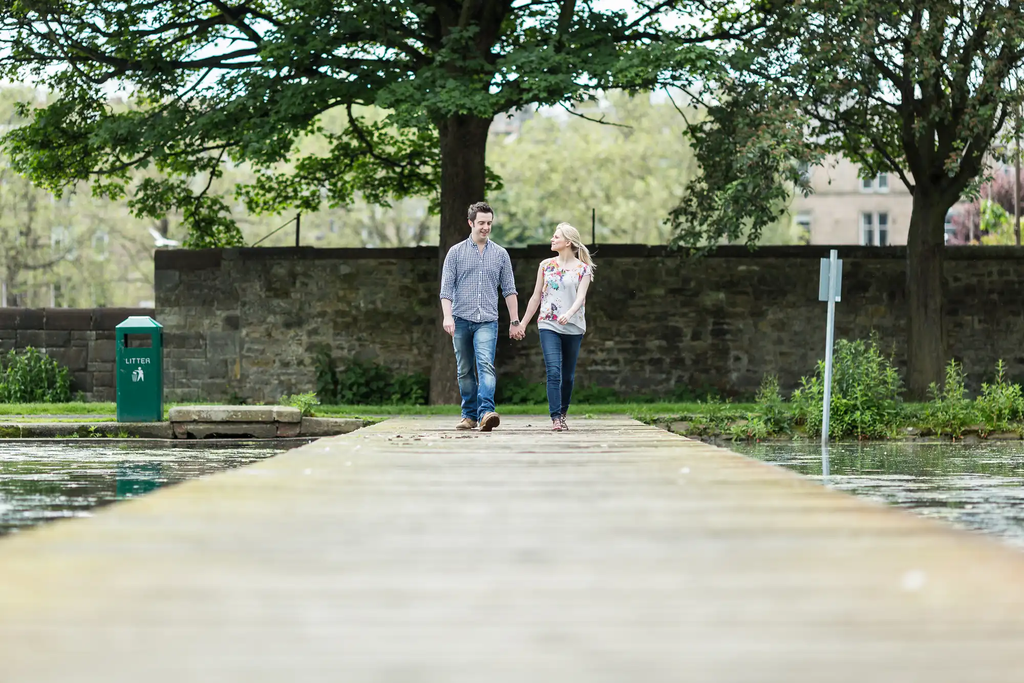 A couple walks hand in hand along a narrow wooden pier in a park, surrounded by trees and a stone wall in the background.