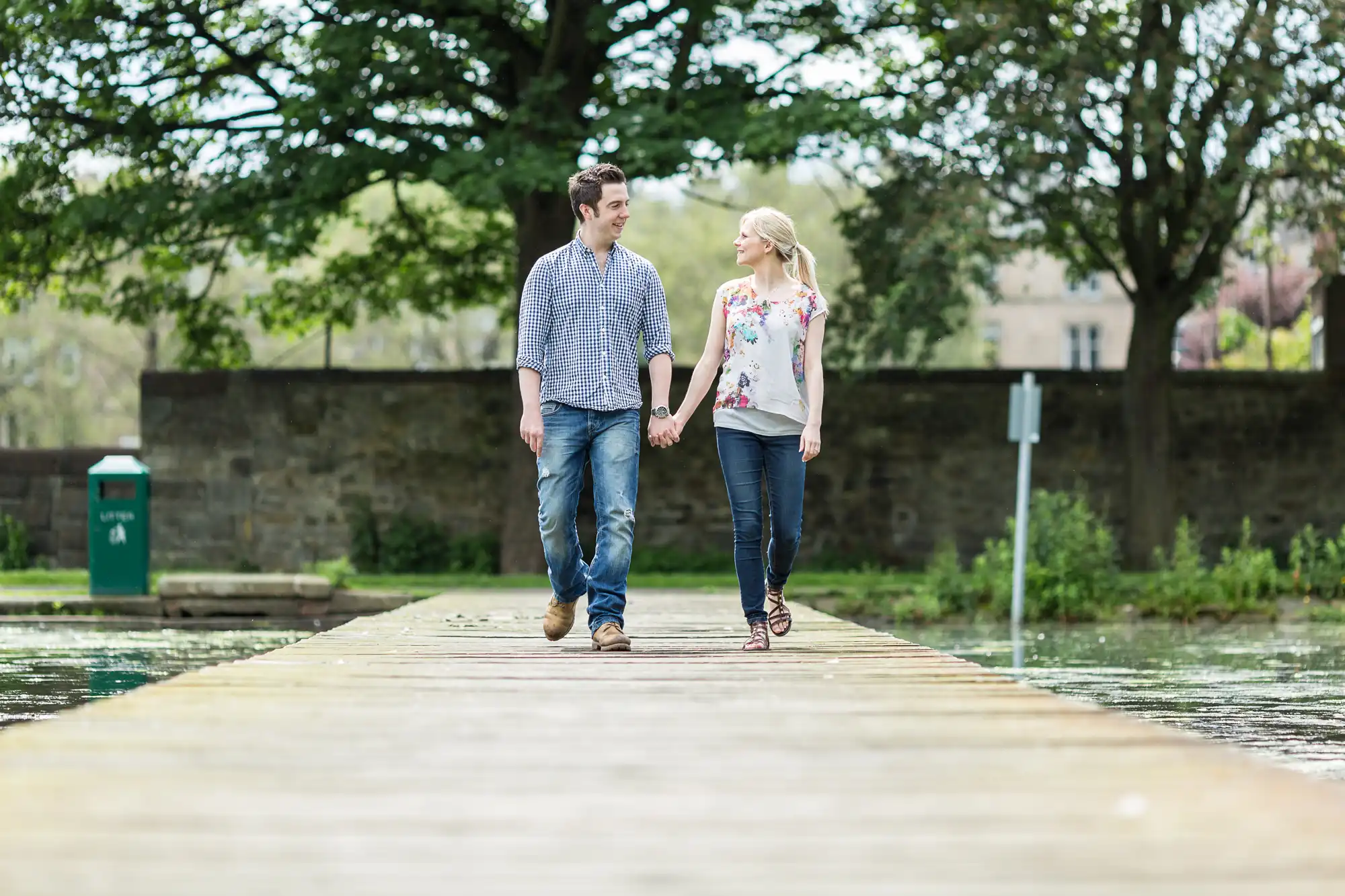A couple holding hands walking down a wooden pier, both smiling at each other, with trees and greenery in the background.