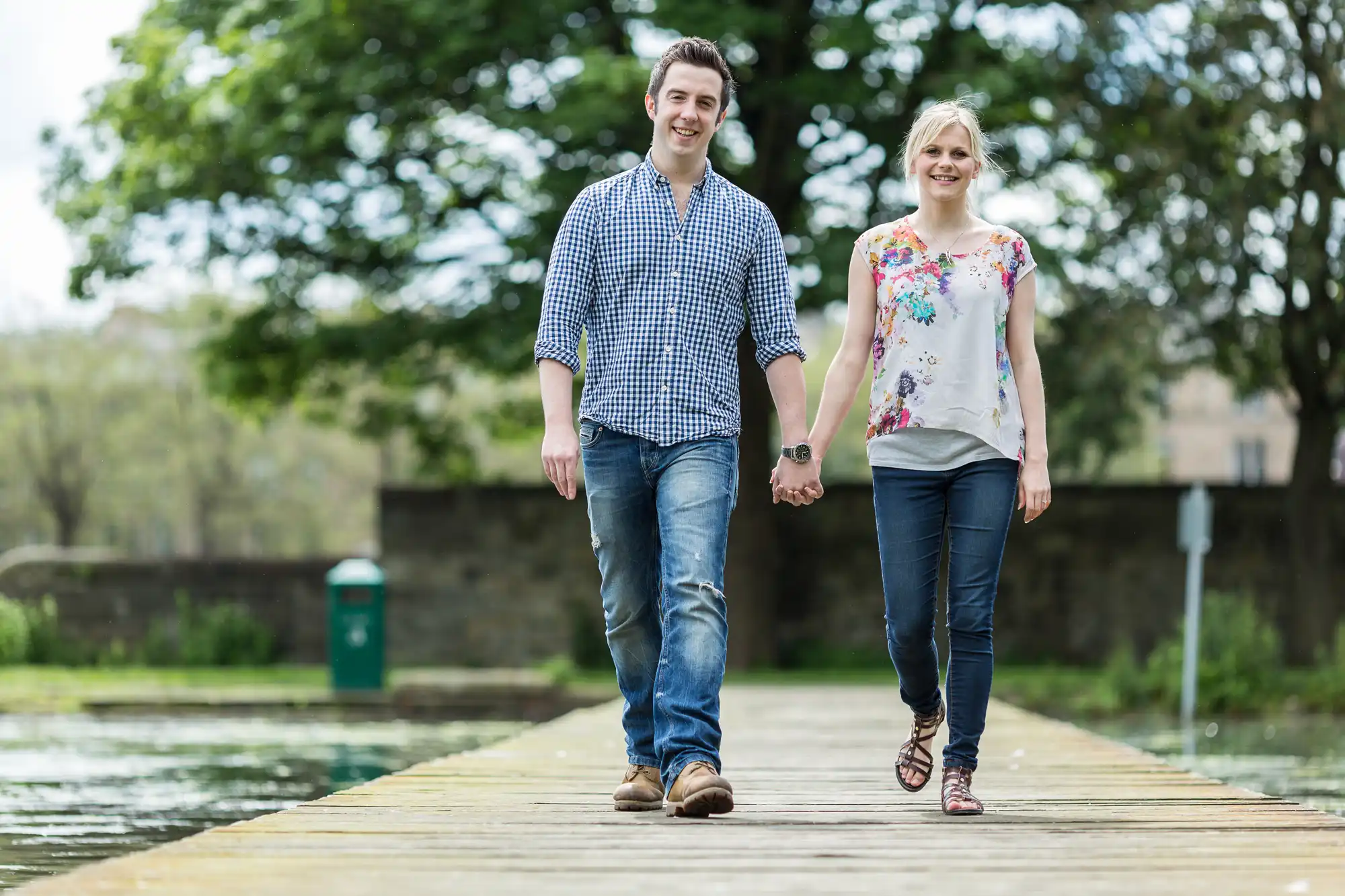 A young couple holding hands and smiling while walking on a wooden dock in a park, surrounded by trees and a pond.