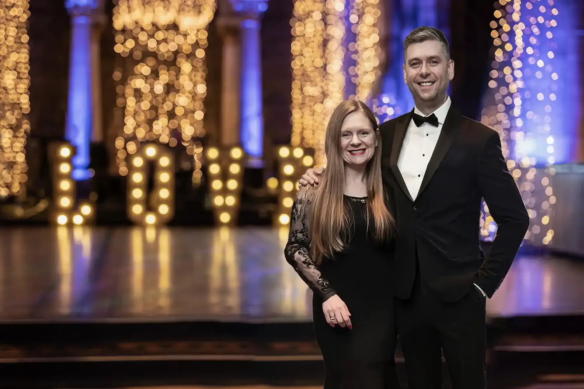 Claire and Jonathon Fowler dressed in formal attire stands in front of an illuminated "LOVE" sign with a background of decorative lights at Mansfield Traquair in Edinburgh