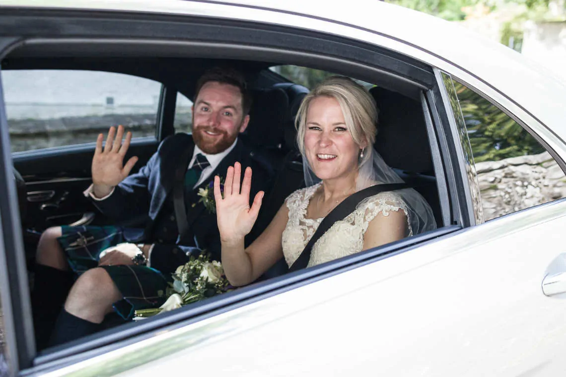Bride and groom wave goodbye from inside the wedding car