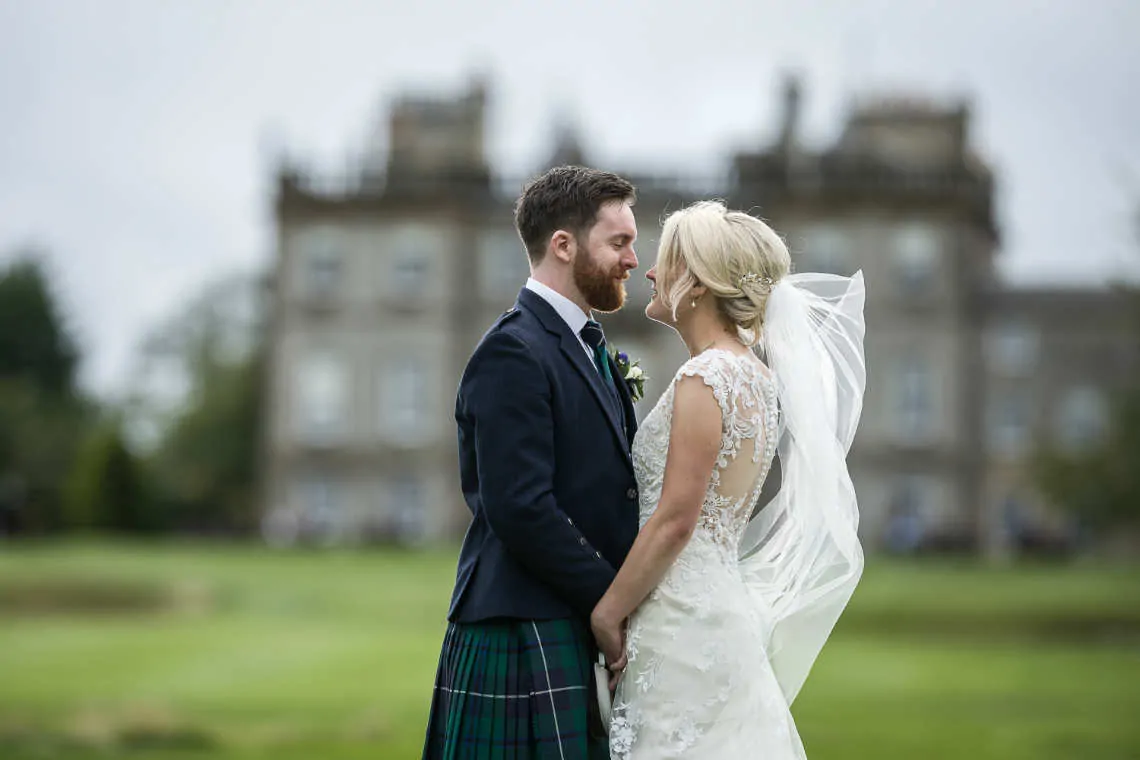 Newlyweds facing each other holding hands with blurred Dalmahoy Hotel in the background
