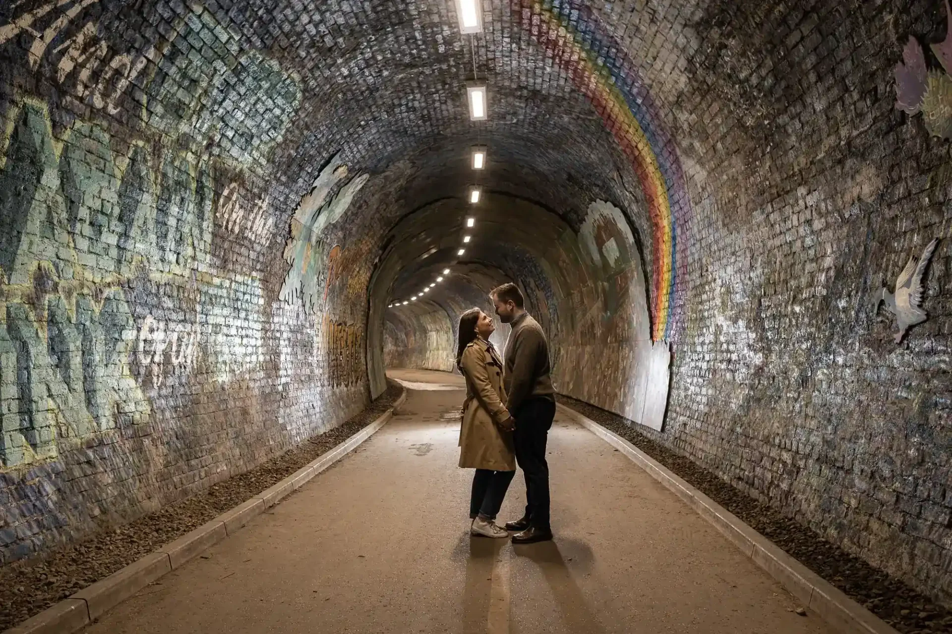 A couple stands face to face in the middle of a mural-covered tunnel with a rainbow and animal paintings on the brick walls under overhead lights.