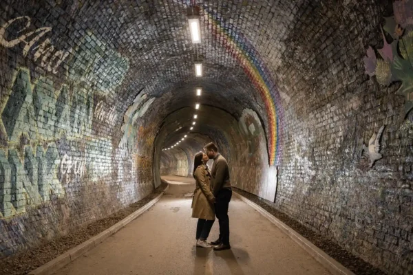 Guided tour of the best photo locations in Edinburgh for wedding and engagement shoots. A couple stands close together in the centre of a brick tunnel decorated with murals, including a rainbow and birds, under overhead lights.