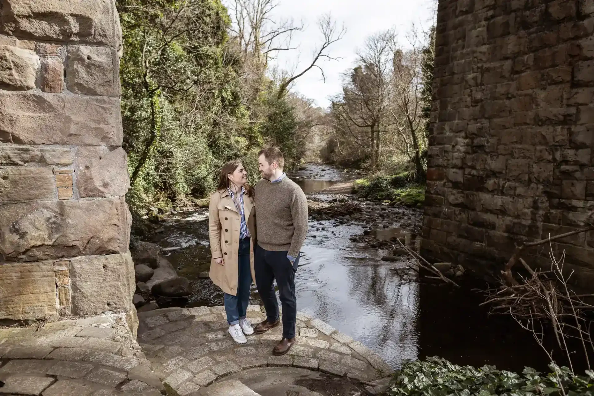 A man and a woman stand facing each other on a stone path next to a river, with trees and large stone structures on either side.