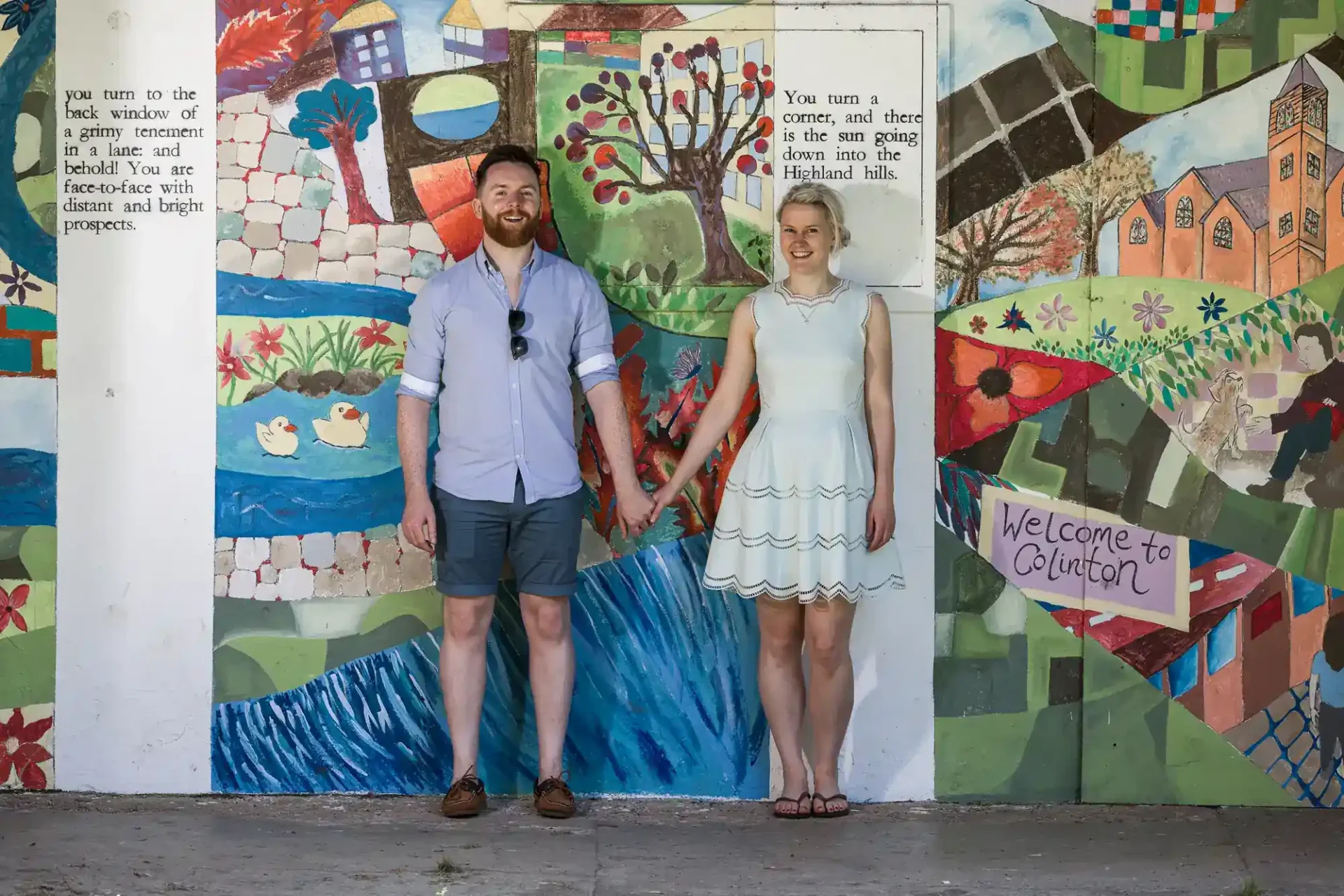 A man and woman hold hands and stand in front of a colorful mural featuring houses, trees, ducks, and a "Welcome to Govilon" sign.