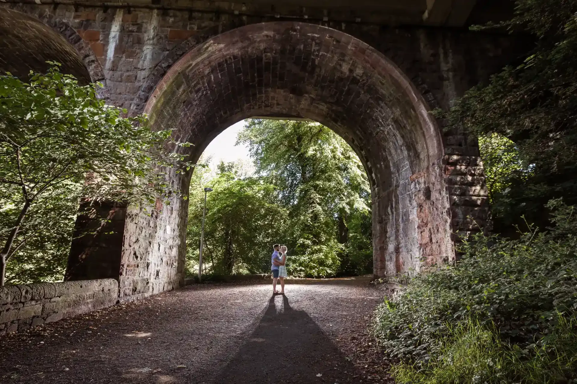 A person stands under a large stone archway surrounded by trees, with sunlight streaming through the opening.