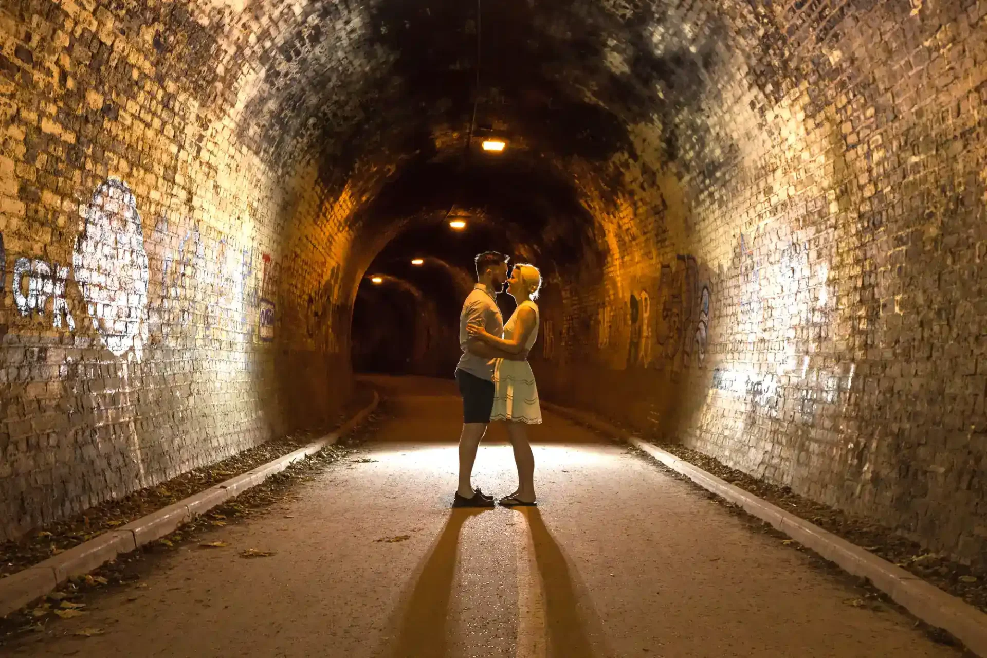 A couple stands embracing and facing each other in the center of a dimly lit tunnel with brick walls and graffiti.