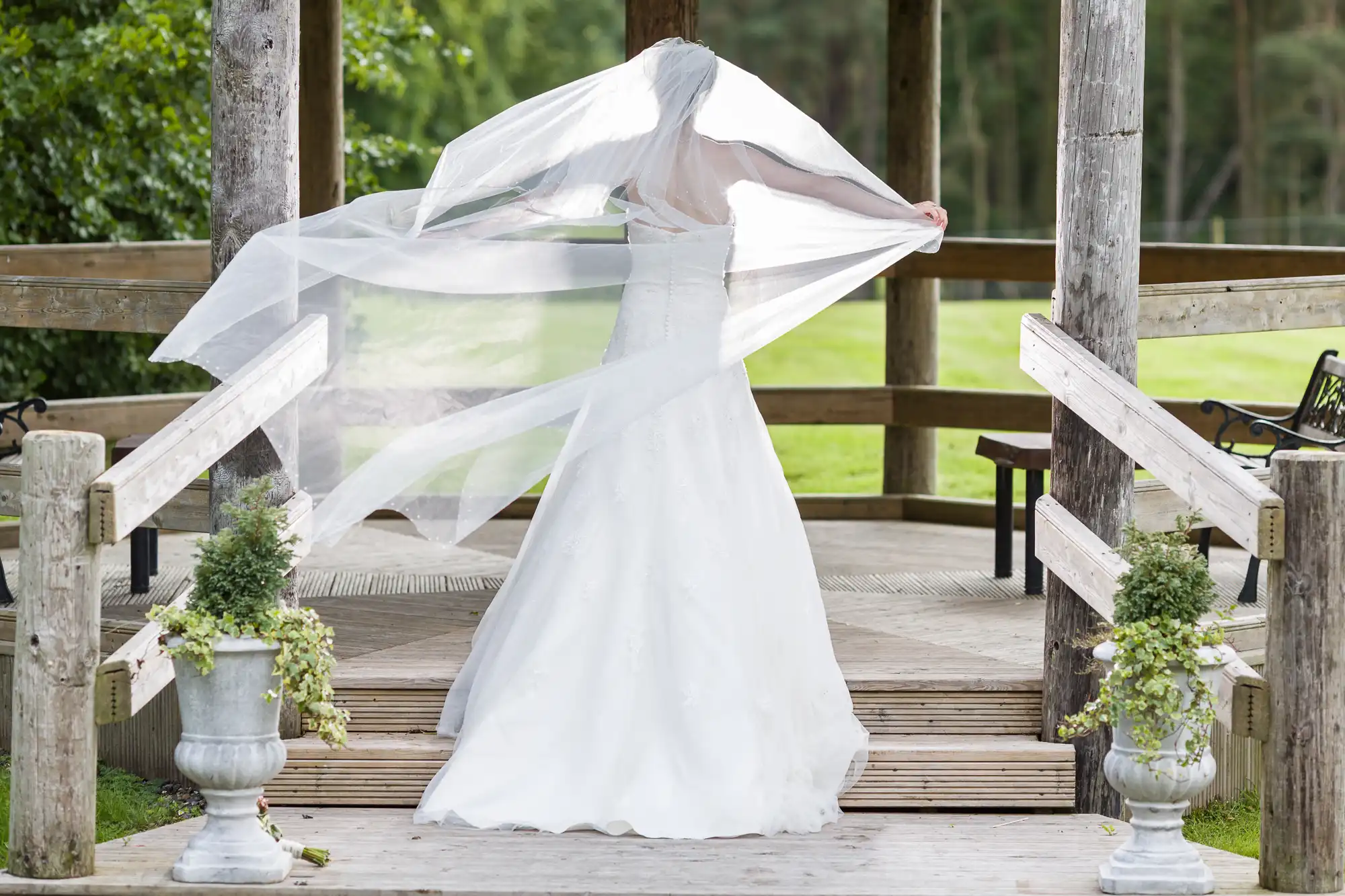 Bride in a white wedding gown holds her veil out, standing on a wooden platform surrounded by greenery.