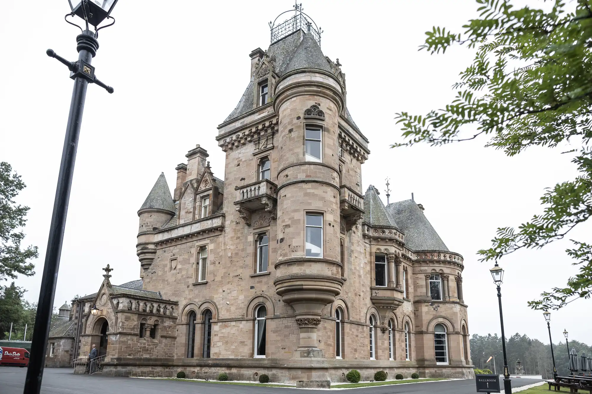 A large stone castle with multiple turrets and ornate architectural details, surrounded by green trees and lamp posts on a cloudy day.