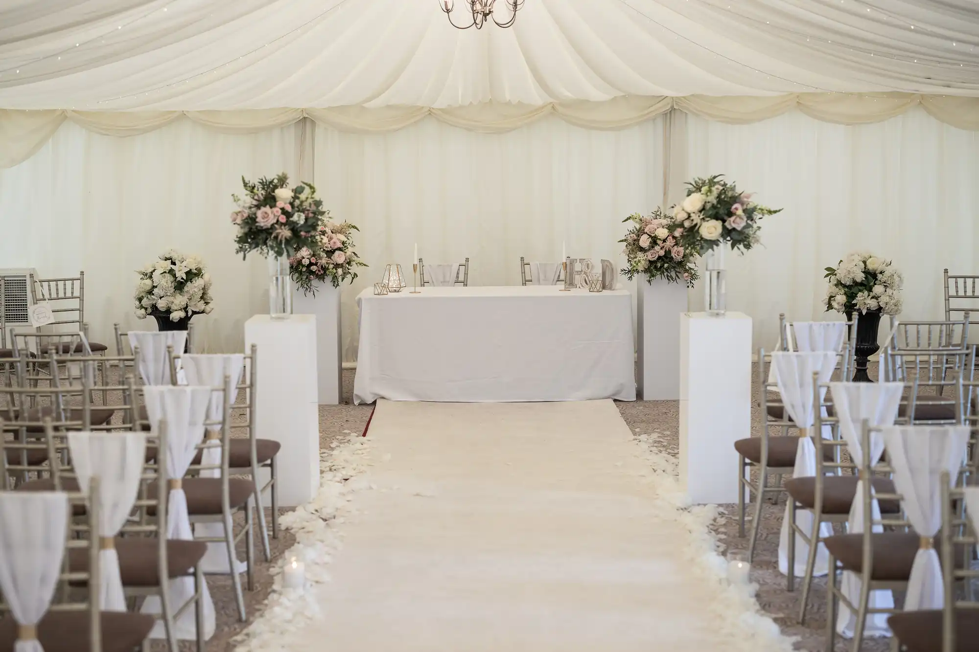 An indoor wedding venue with white drapery, arranged chairs, floral decorations, and a central aisle leading to a table with chairs at the front.