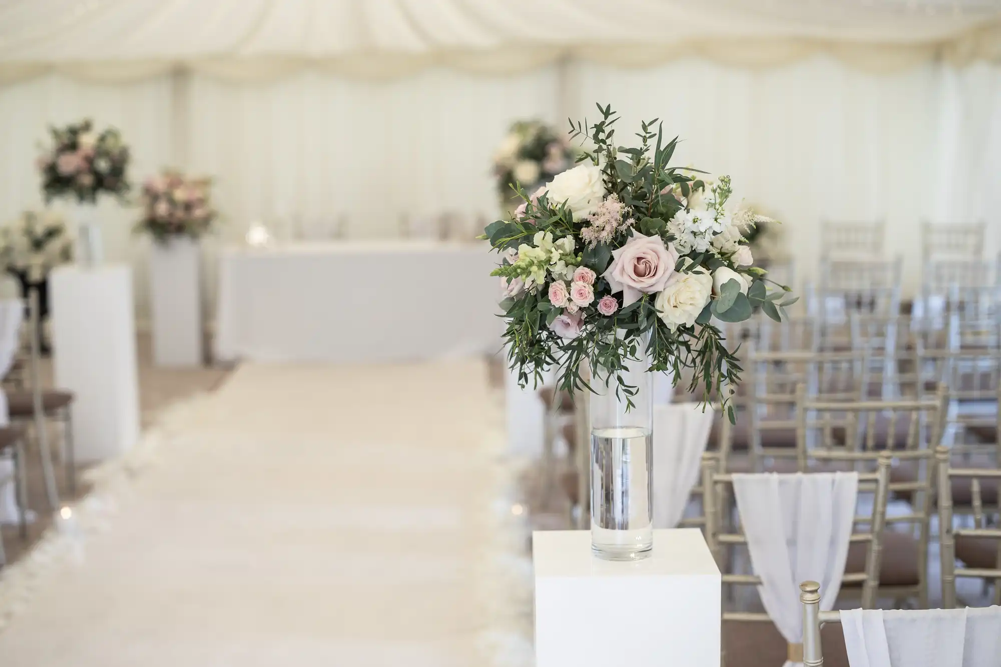 A decorated wedding aisle with white and pink floral arrangements, transparent vases, and rows of gold chairs draped in white covers. A white table is visible at the end of the aisle.