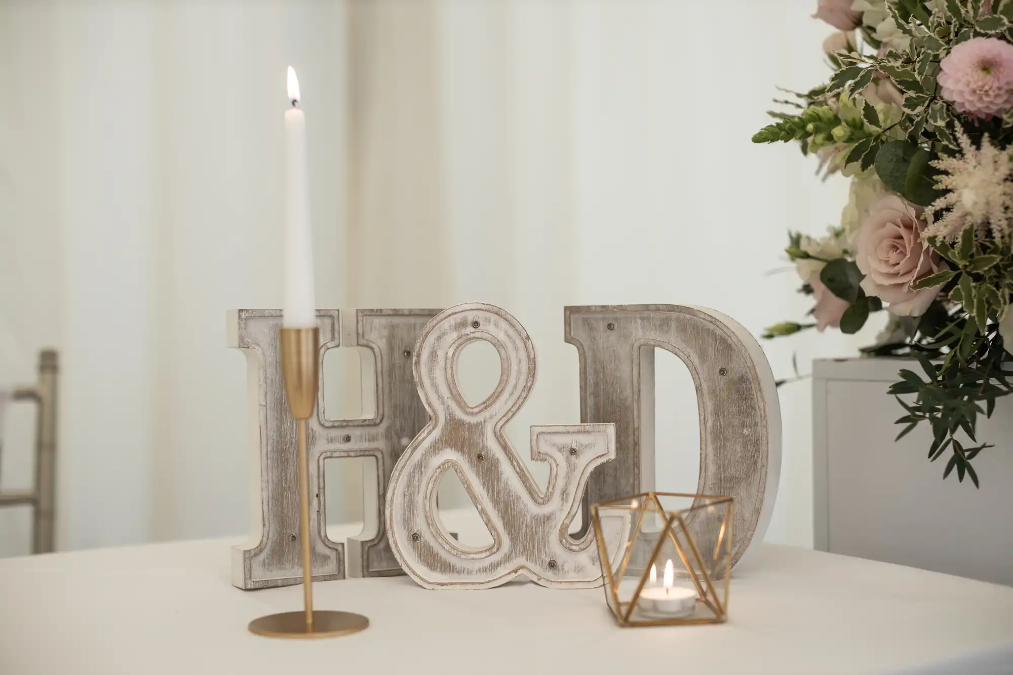 Wooden letters "H & D" are displayed on a table with a lit candle in a gold holder, another lit candle in a geometric holder, and a floral arrangement in the background.